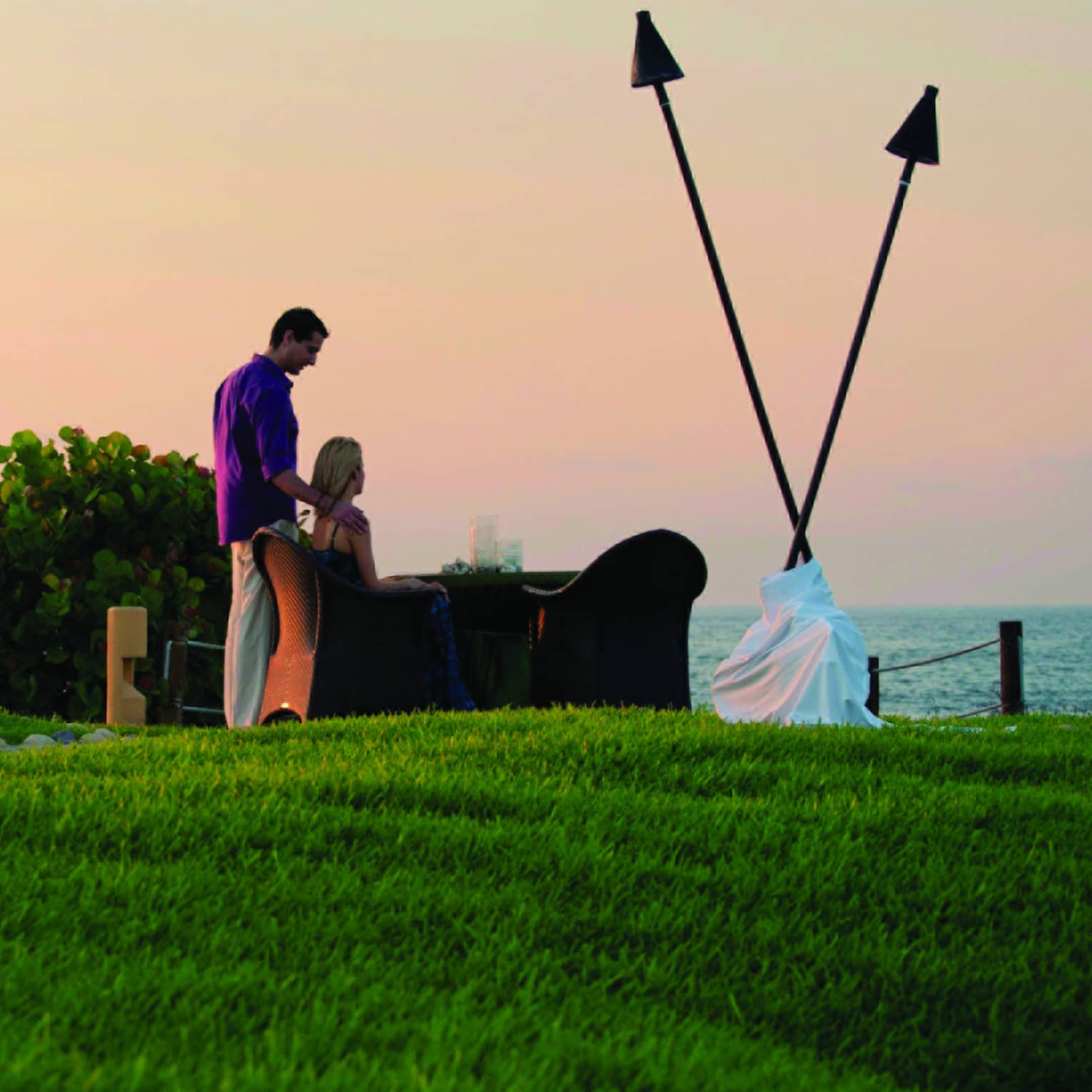 On a grassy hill with a huge arrowhead artwork, a couple at a table gaze at a calm ocean and hazy orange sky on the horizon. 