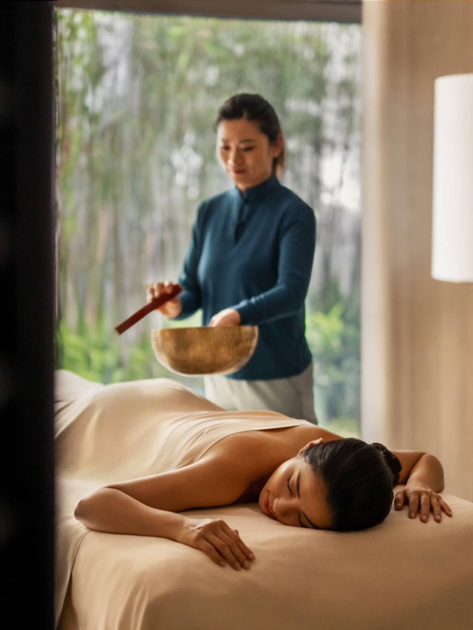 Female spa therapist performs singing bowl spa ritual for woman on spa treatment table and luxury hotel spa
