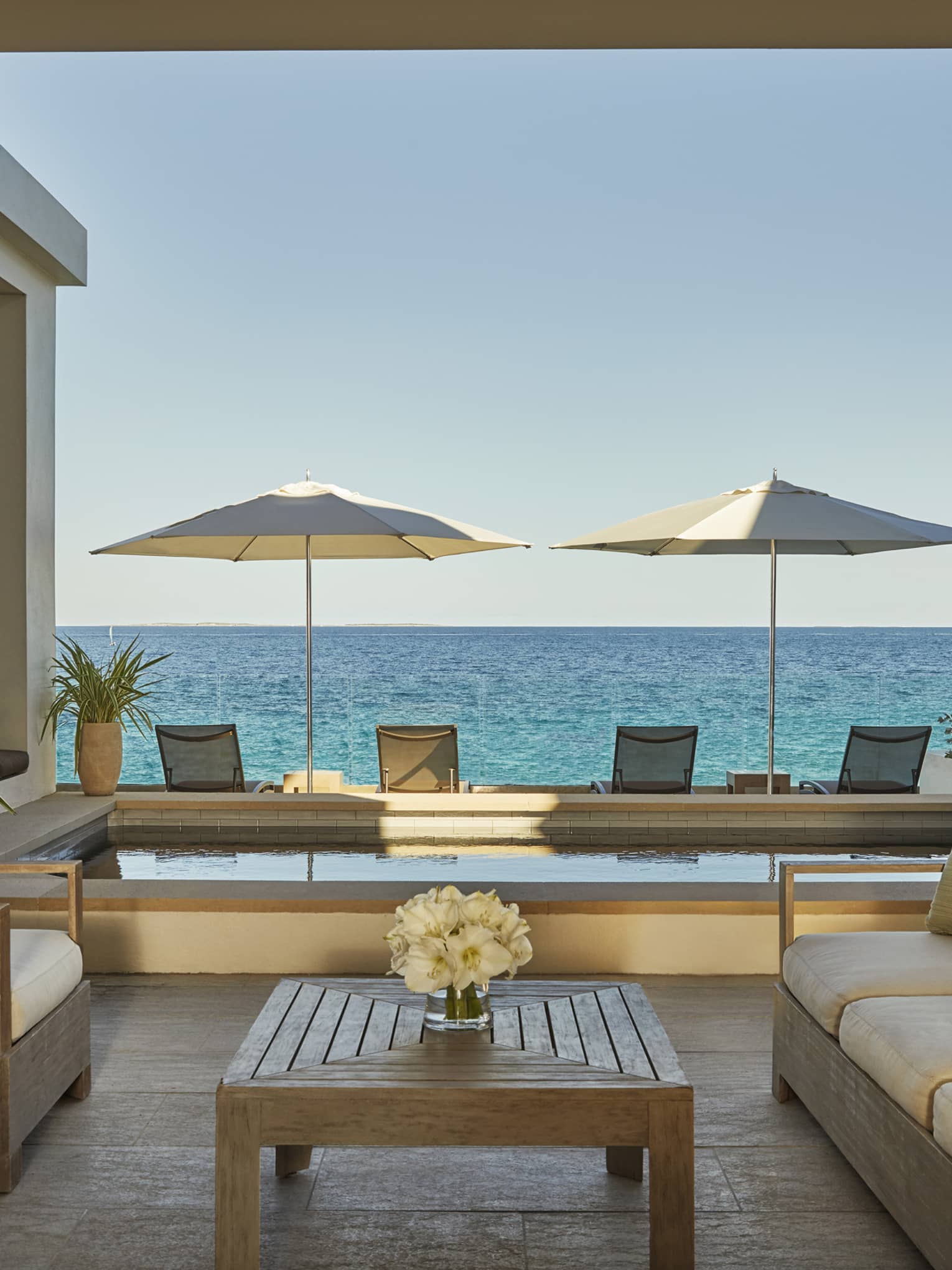 Outdoor terrace with cushioned seating, a wooden table, two umbrellas and ocean views beyond a private pool