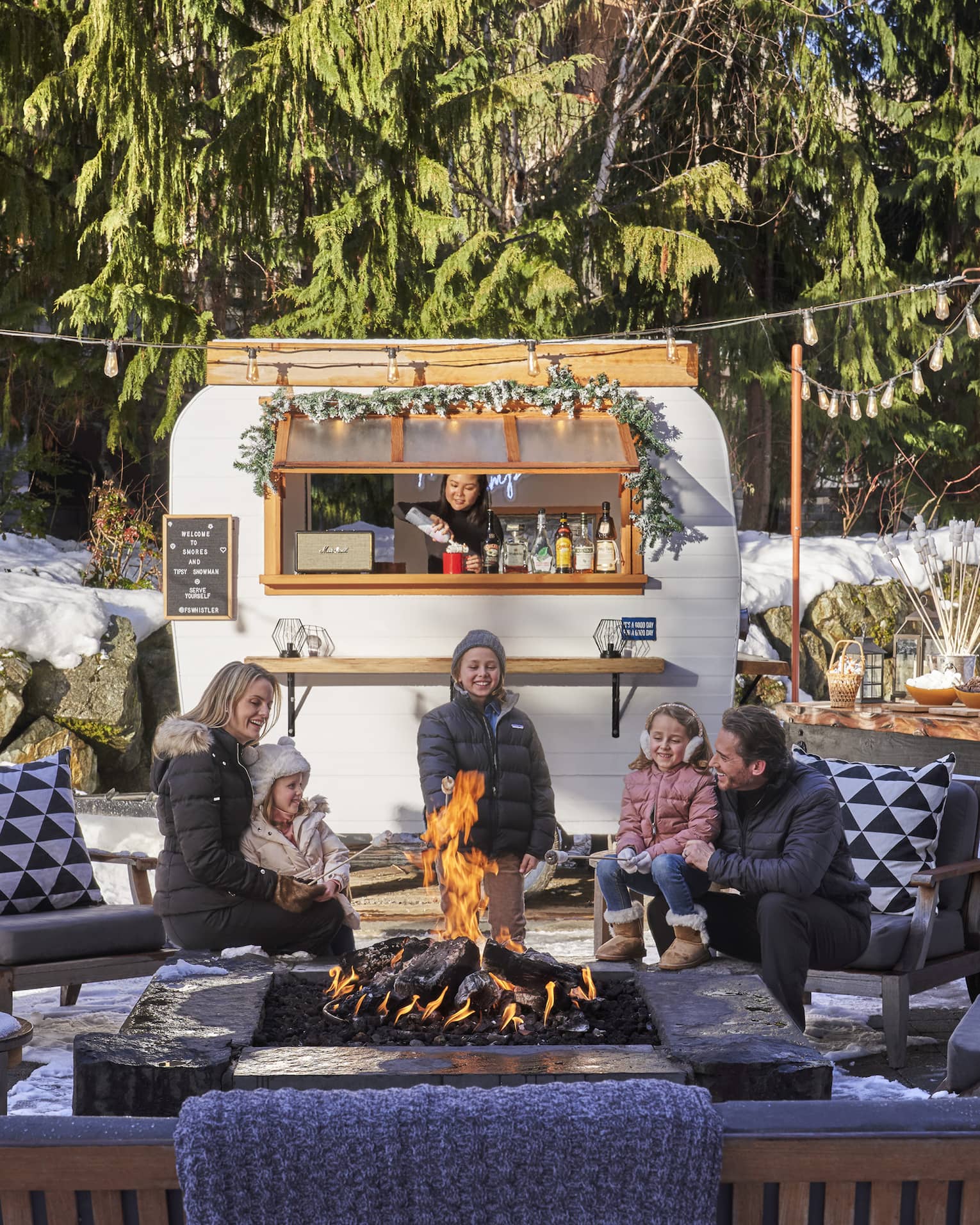 An outdoor camper sits behind a young family as they talk around a fire.