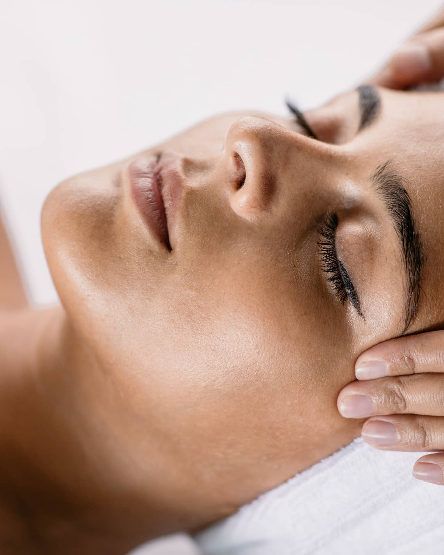 Hands massage woman's temples as she closes her eyes, spa towel around hair