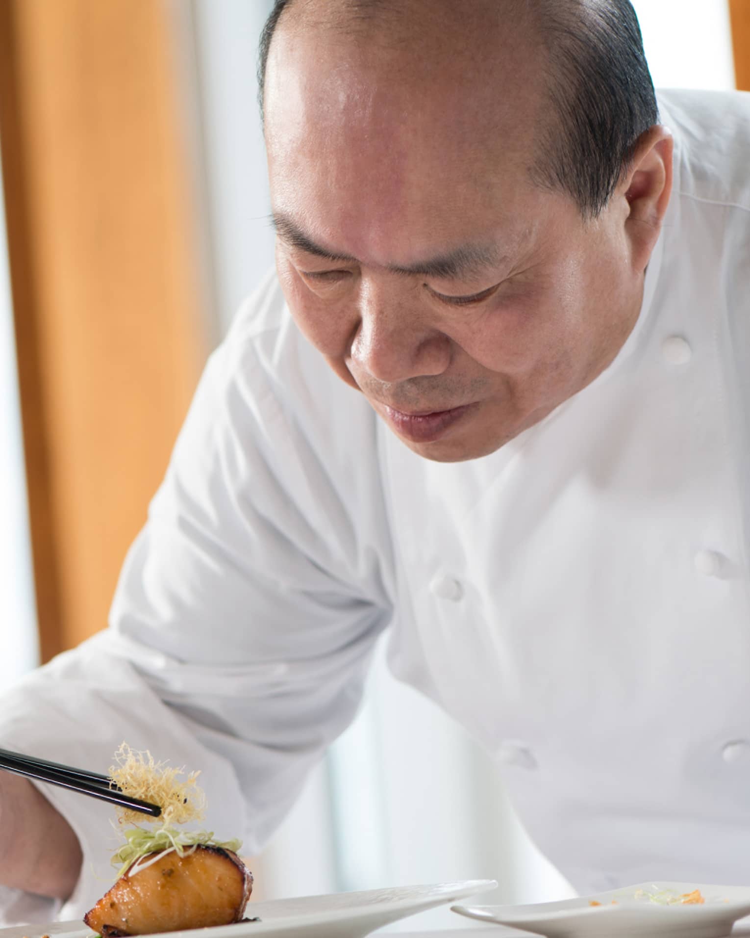 Chef in white uniform leans over grilled salmon dish on plate, adds garnish with chopsticks