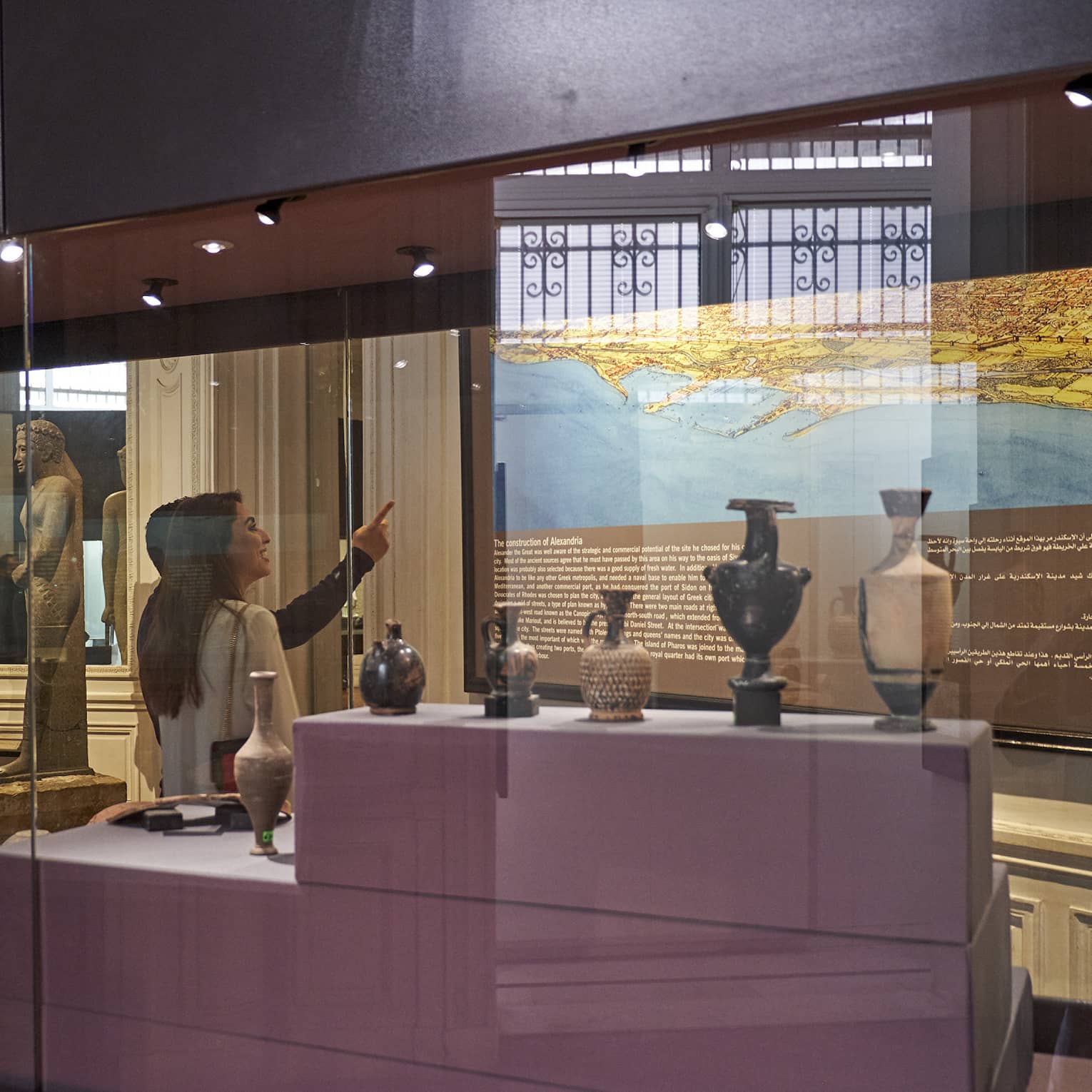 A visitor in a museum points up at a historical map lit up on a wall, behind them a display case with various vases and jugs.