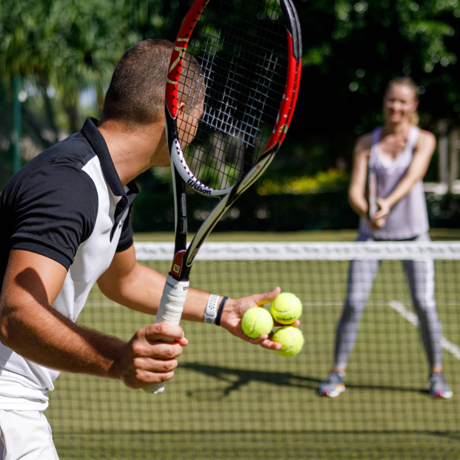 Couple playing tennis on court, man prepares to serve tennis balls to woman ready to swing
