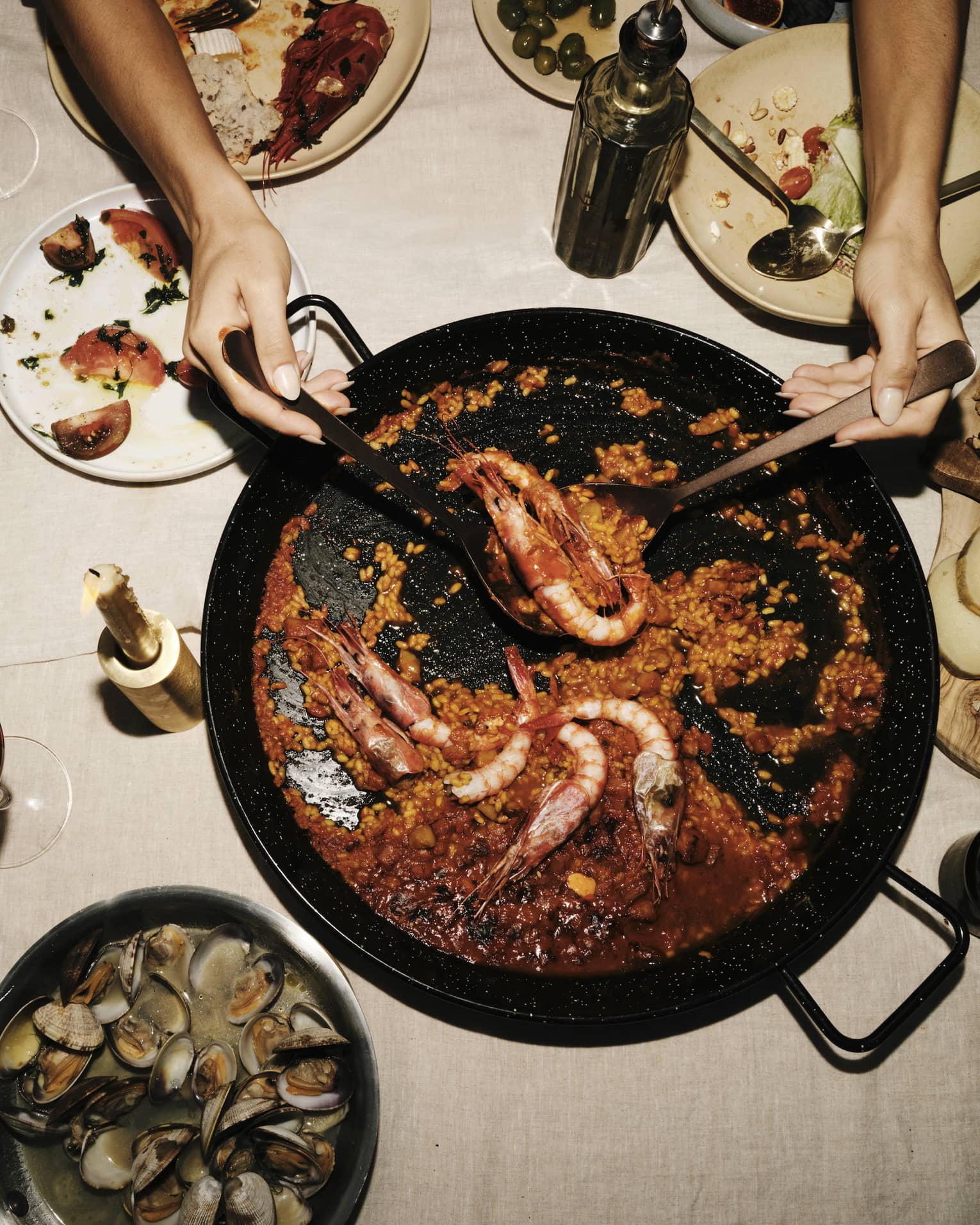 Two hands use large spoons to lift prawns from a saucy paella dish set amid plates of seafood, cheese and fruit.