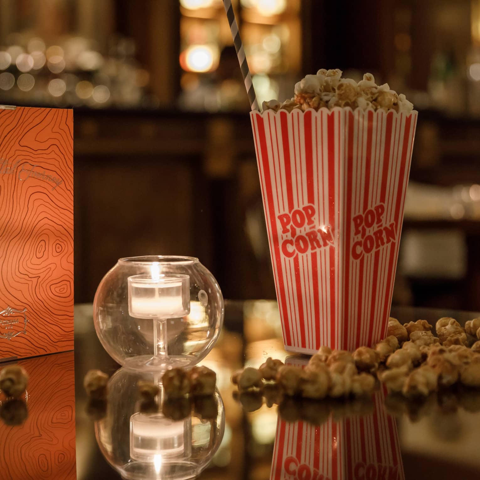A red and gold box of chocolates, a candle in clear glass and a tall box of popcorn on a shiny table reflecting warm lights.