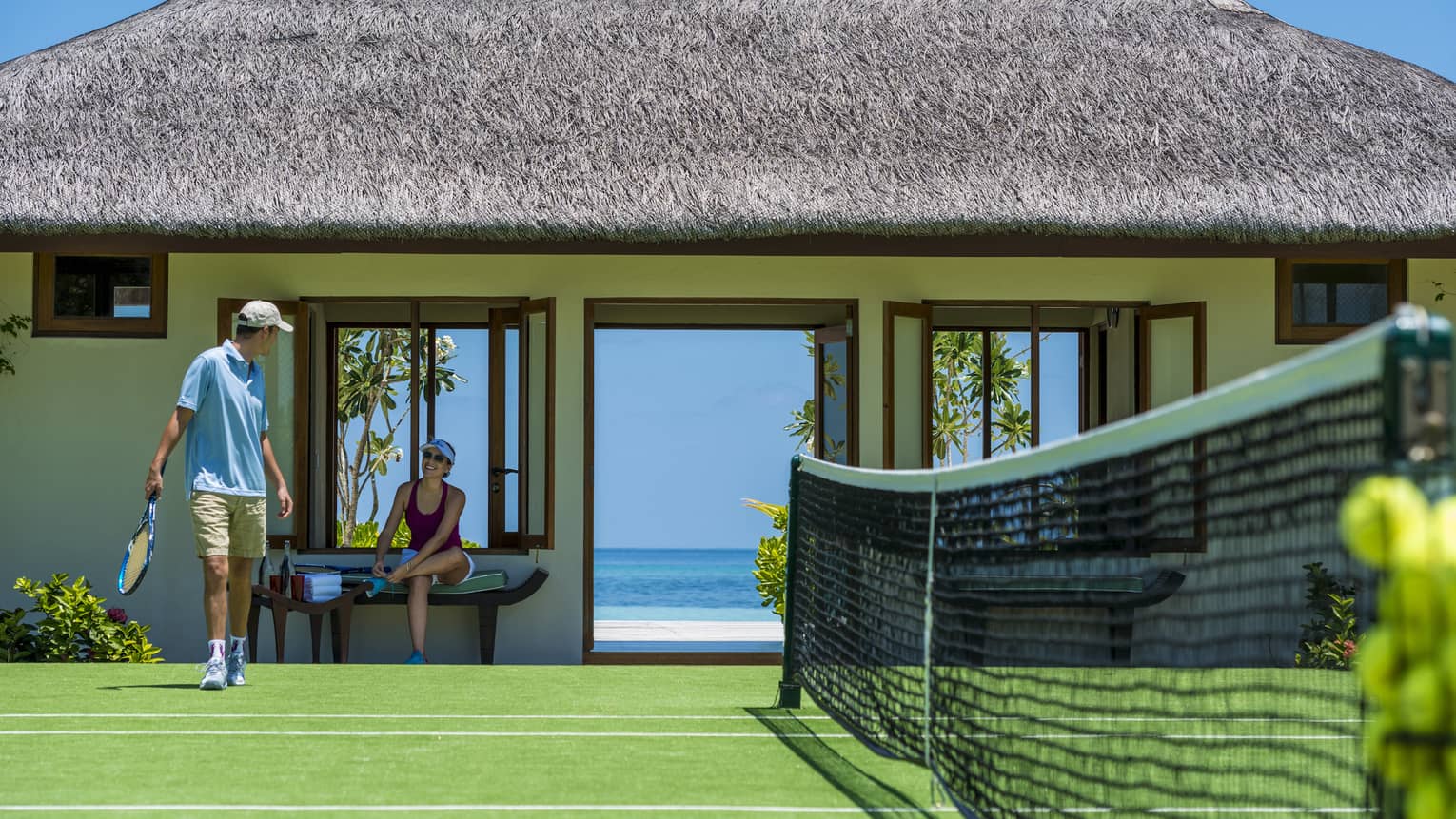 Two guests prepare to play tennis on a court bordered by a grass-roofed building with doors opening onto a view of the ocean.