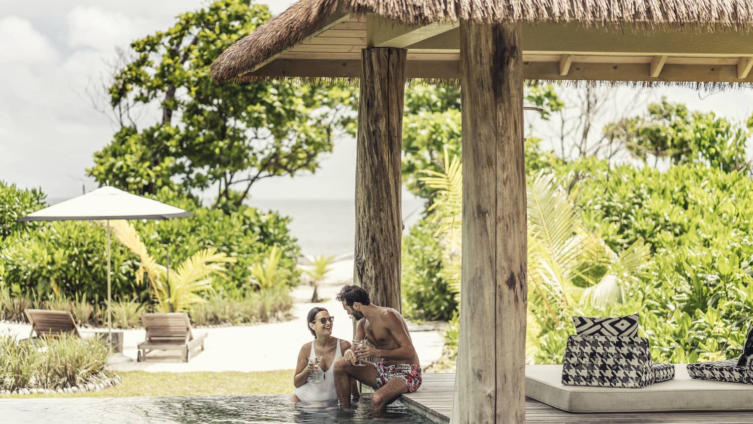 Couple in villa pool under thatched roof and view of tropical foliage