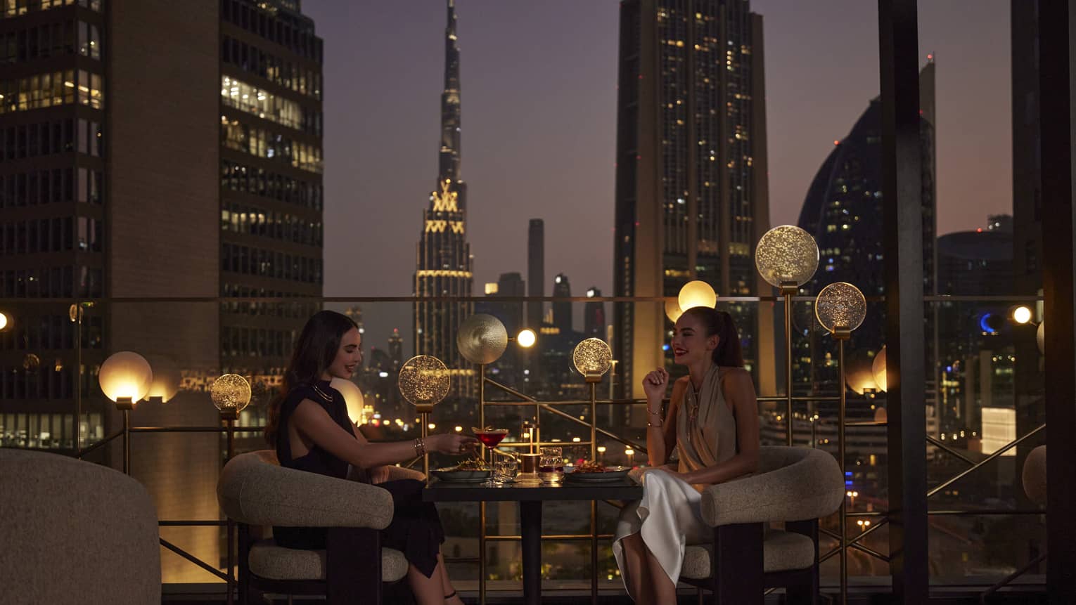 Two women sit at a cocktail table on an outdoor rooftop patio overlooking the Dubai skyline at night