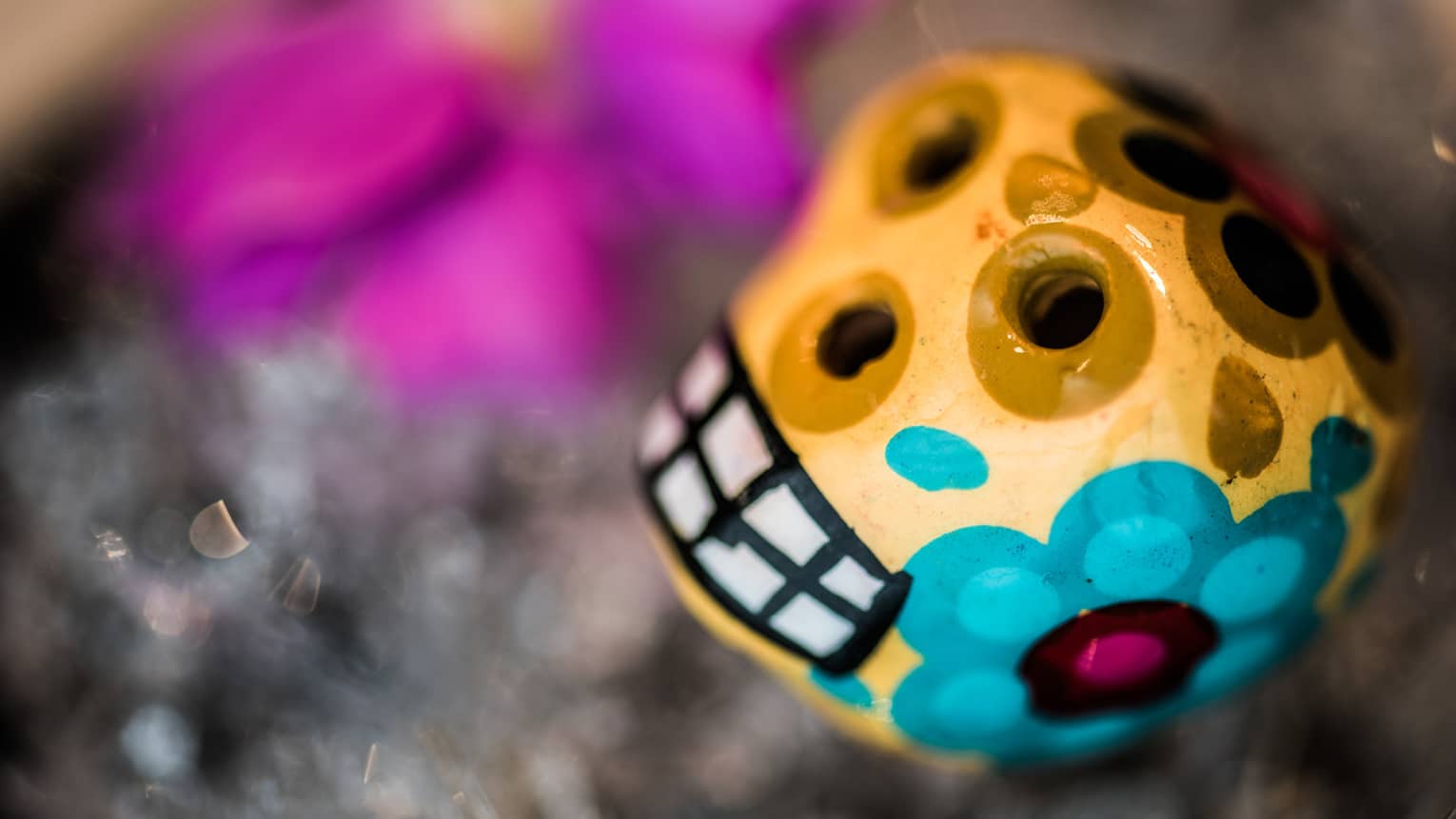 Extreme close-up of a yellow, hand-painted clay sugar skull, featuring large blue and brown flowers with fuchsia centres..