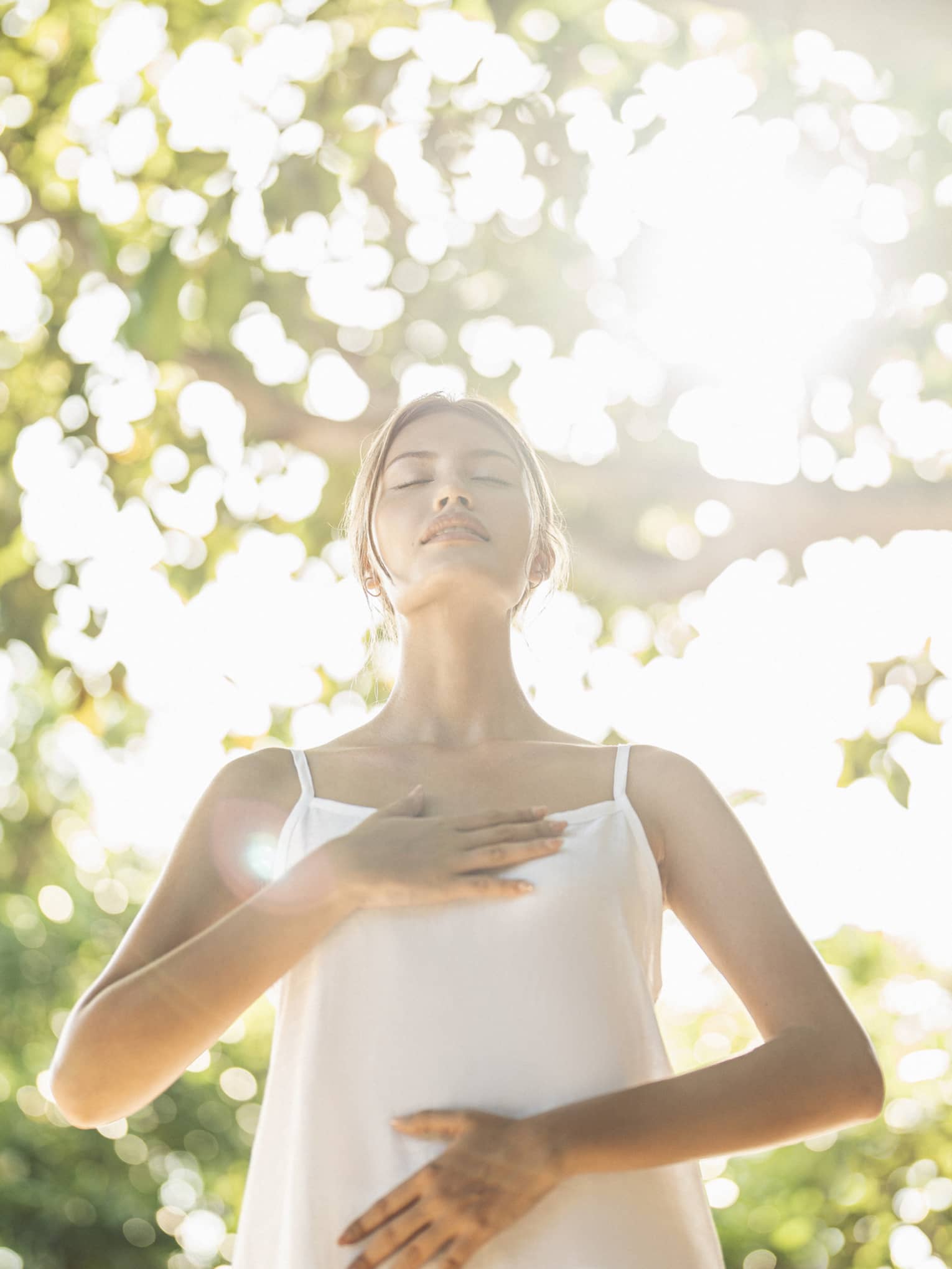 Person practicing mindful breathing in a wellness haven, standing under sunlit trees with hands on chest and abdomen