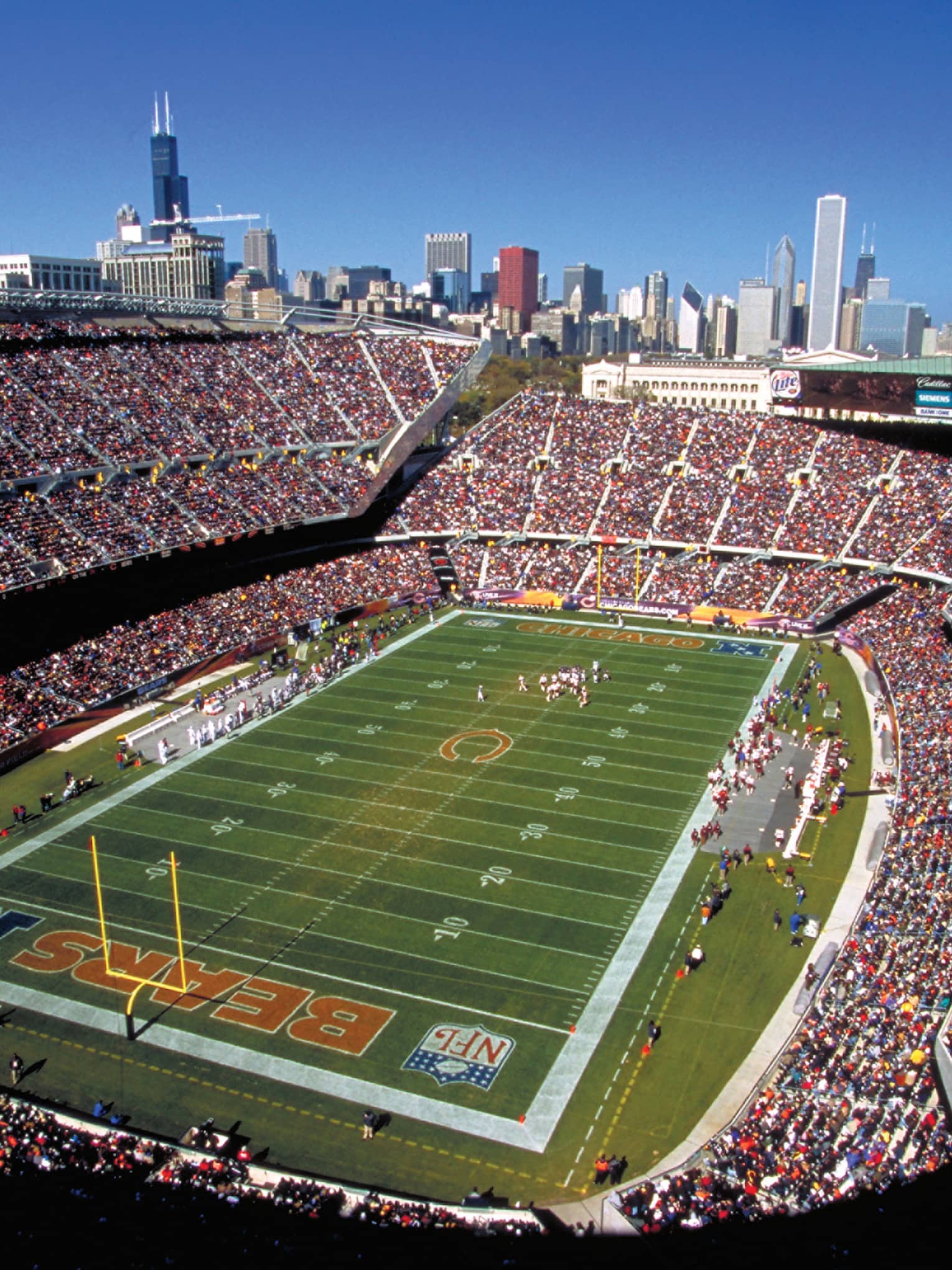 Aerial view of open stadium with football field, stands filled with fans against blue sky