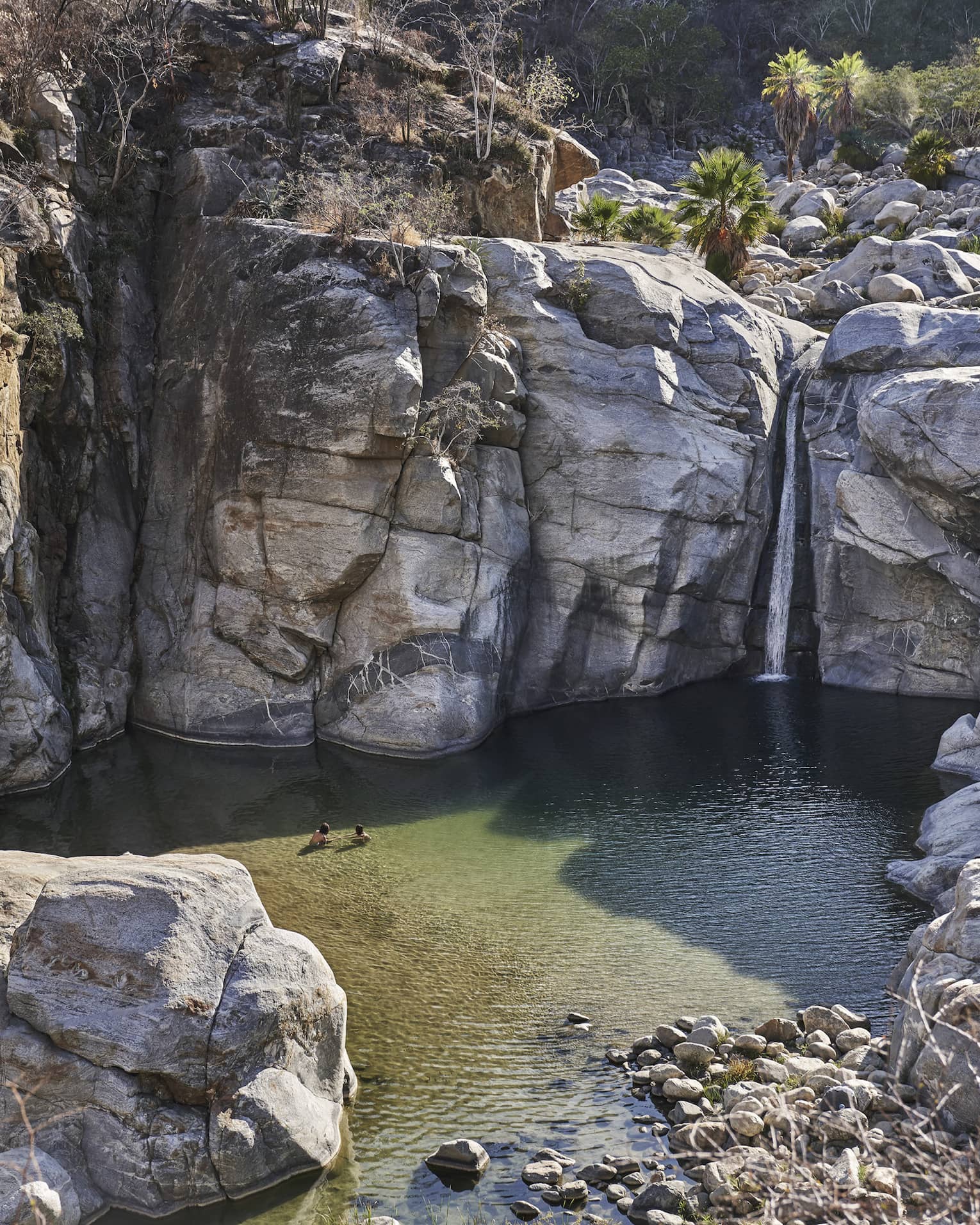 Two people wade in small ocean lagoon under large cliffs