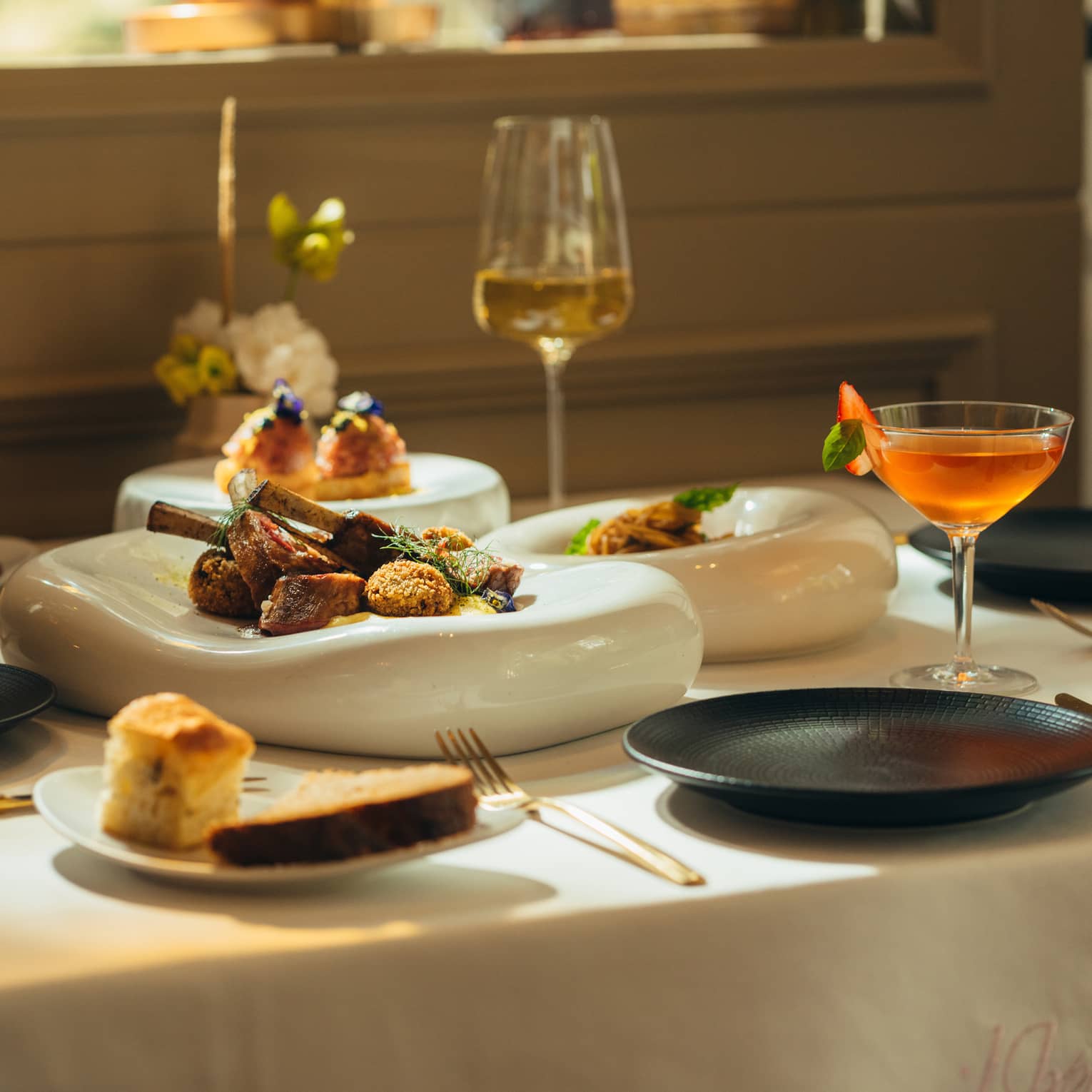 Three white serving platters with food sit in the middle of a white-cloth dining table with three black plates, a glass of white wine, a cocktail in a coupe and two small plates of bread surrounding them