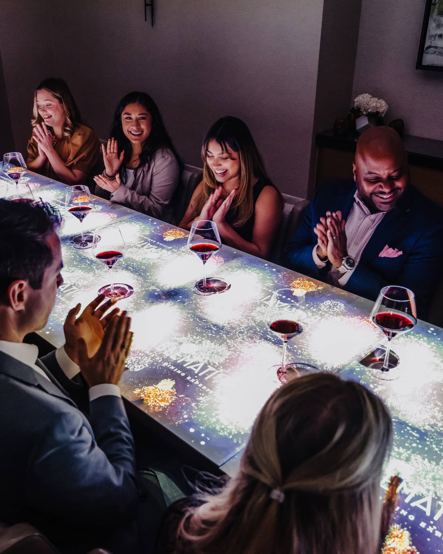 A large group of people sitting at a table with a screen on top of it drinking wine.