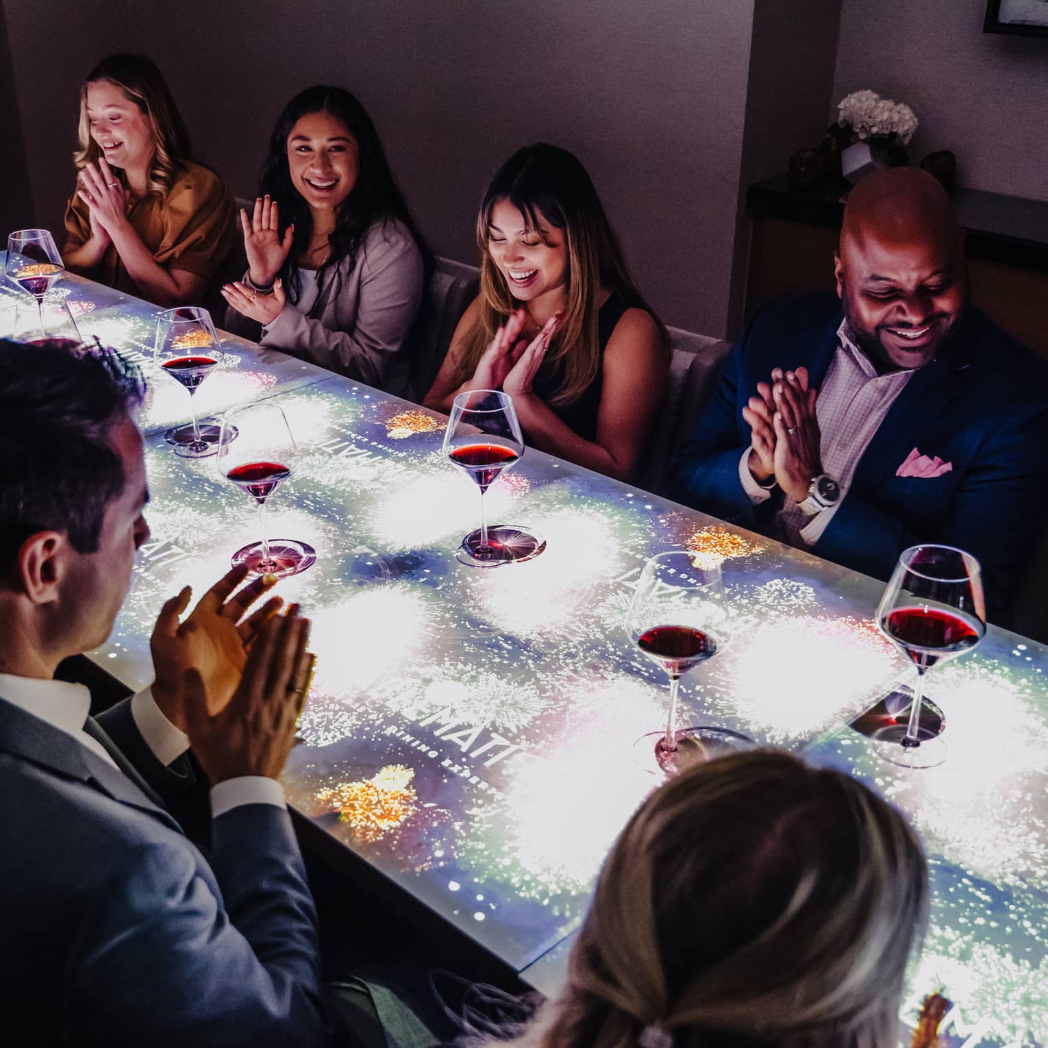 A large group of people sitting at a table with a screen on top of it drinking wine.