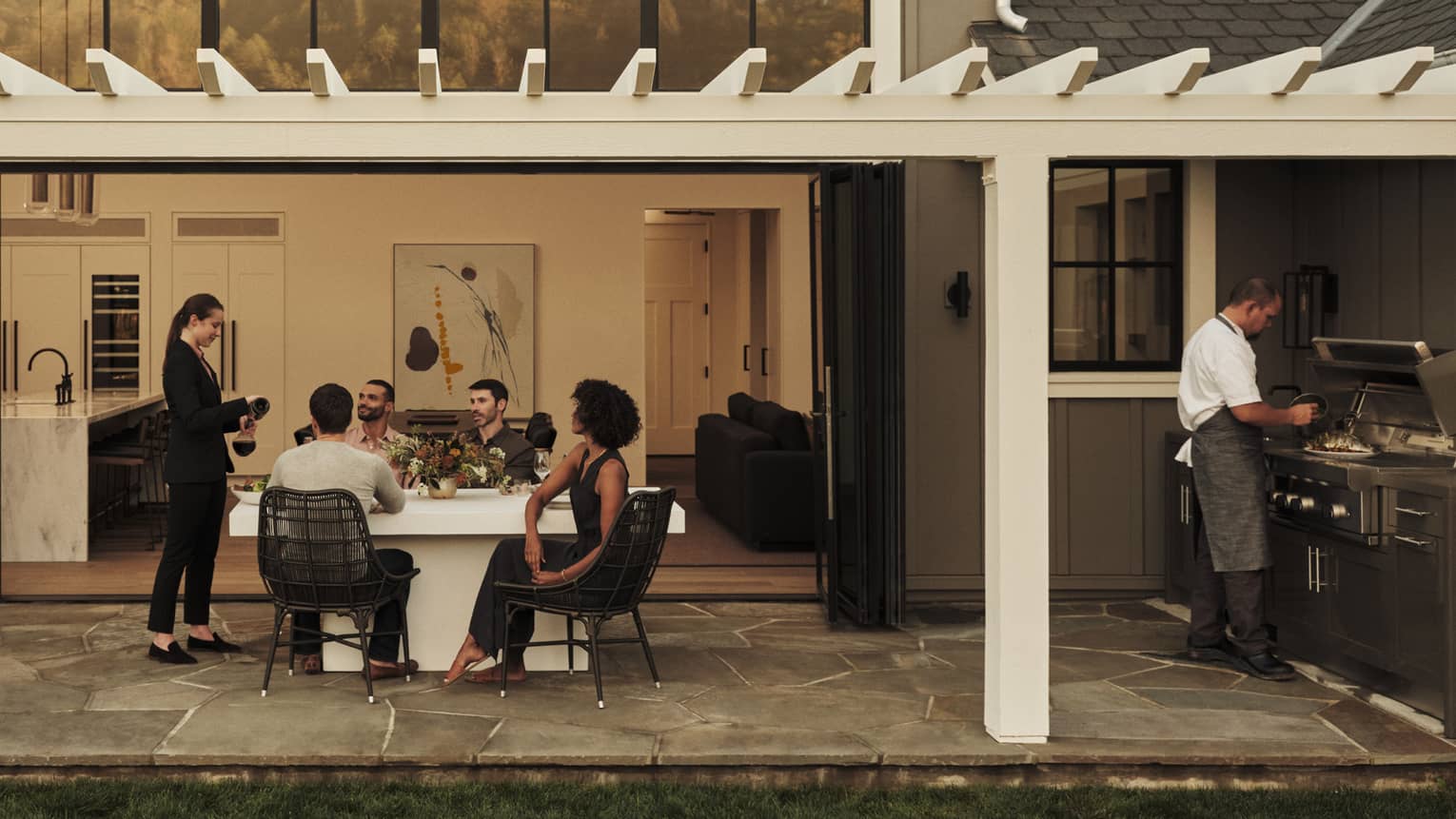 Four guests sit at an outdoor table and watch a server decant a bottle of wine; a chef prepares food at a barbecue nearby.