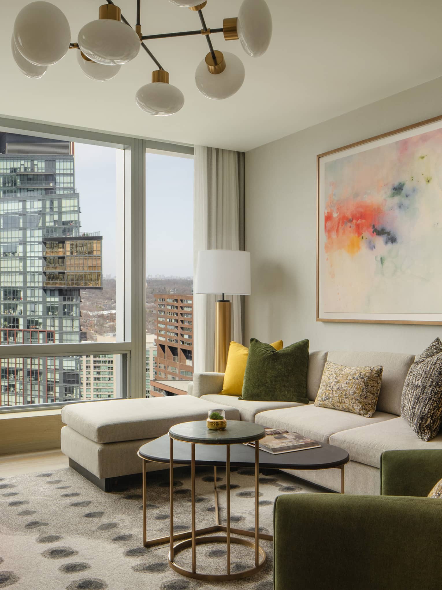 Stylish suite living area with a large sectional sofa, colorful pillows, modern artwork, and floor-to-ceiling windows overlooking the city