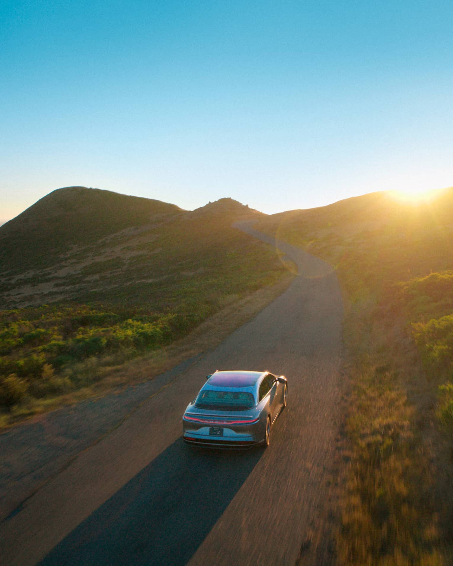 A car drives down a road surrounded by green hilly terrain. A clear blue sky with the sun just peeking over the hills is in the background, and there is also a glimpse of the ocean behind the hills.