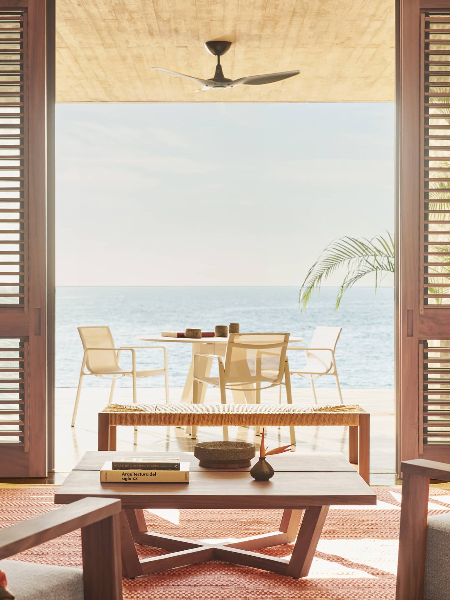 A cozy living room with warm orange accents, featuring a wooden coffee table and a grey sofa. The space opens to a sunlit terrace with ocean views and an outdoor dining area.