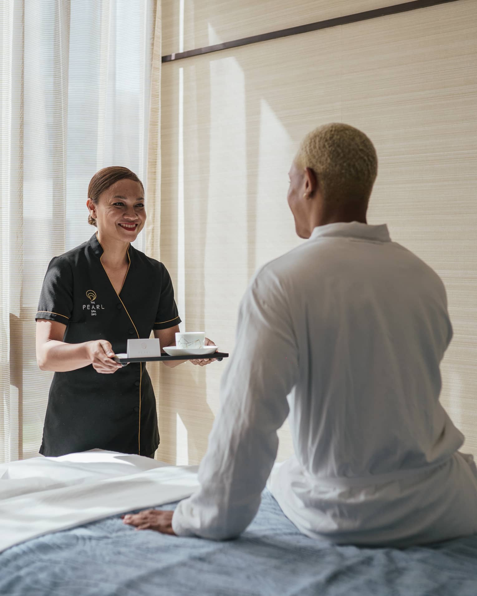 Spa attendant carrying a tea tray greets a man wearing a white spa robe who is sitting on a massage table
