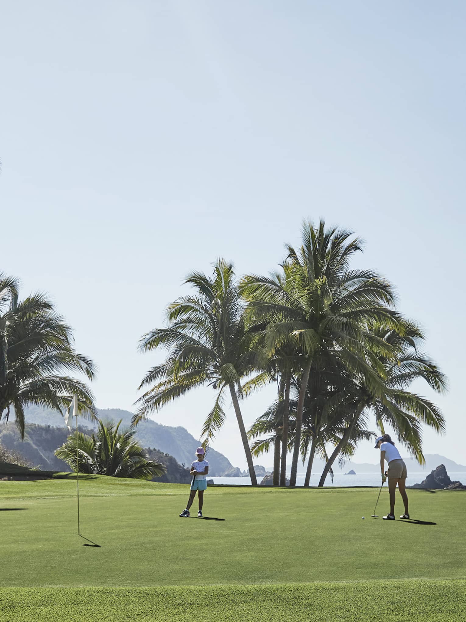 A golfer about to tap a ball toward a nearby hole as a child looks on, on a pristine green bordered by ocean and palm trees.