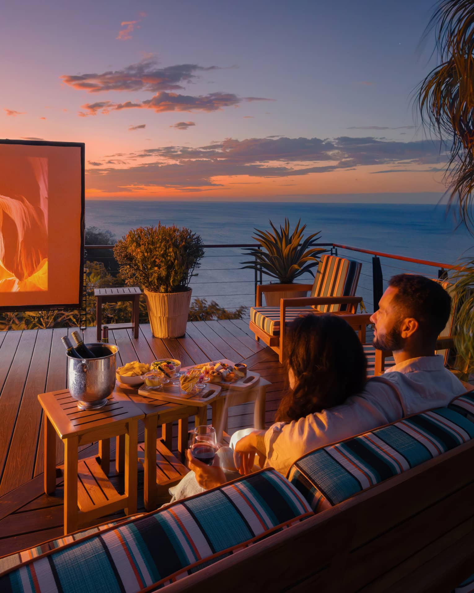 Two people sit on an outdoor sofa with a small table of food infront of them as they watch a movie on a large outdoor screen set up on a private terrace