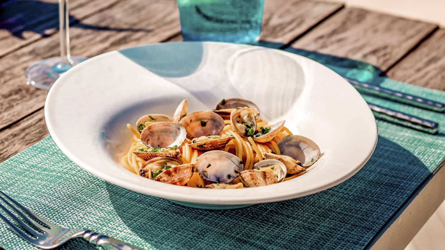 Spaghetti alle vongole on wooden picnic table with turquoise placemat and water glass, clear wine glass, conch shell