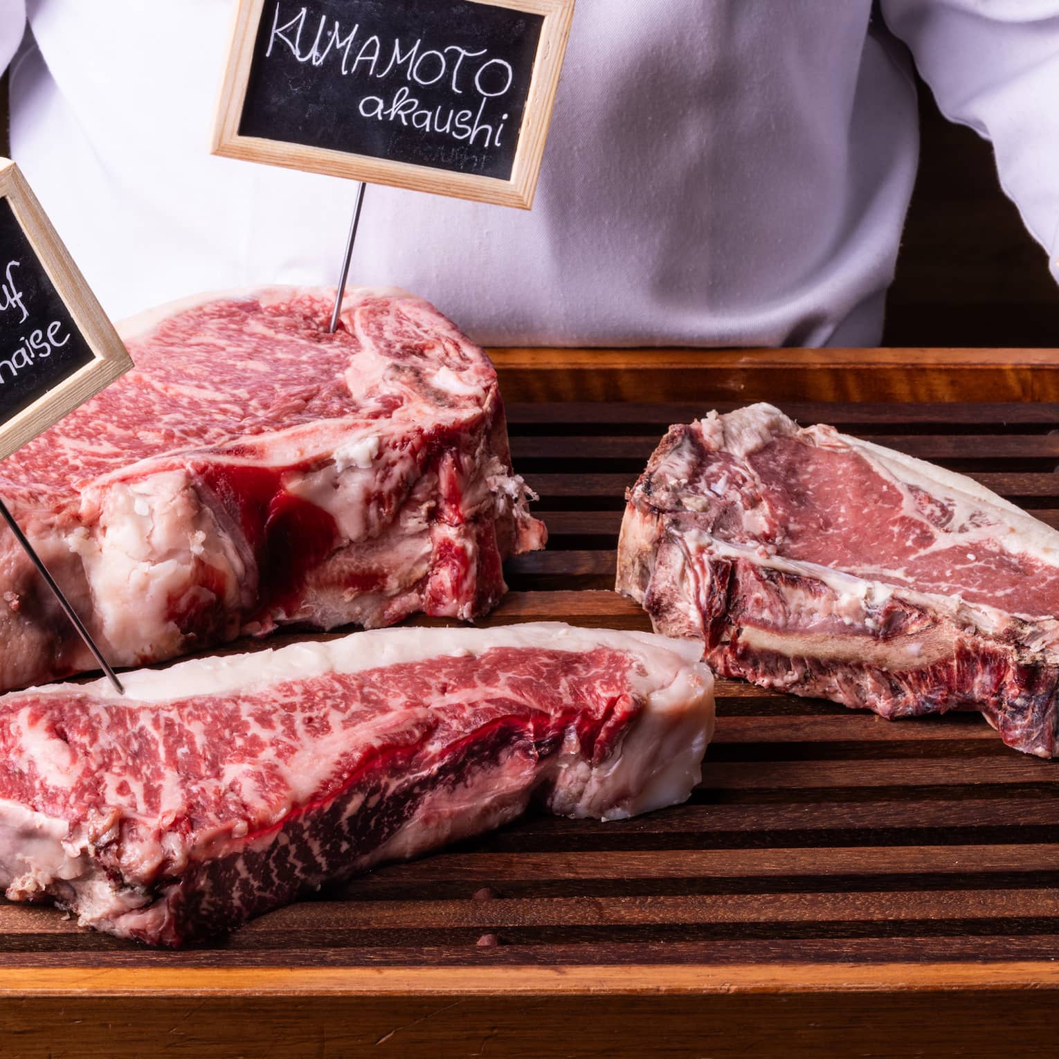 Close-up view of a wood tray with three labelled cuts of well-marbled beef, held by person in white chef's coat.