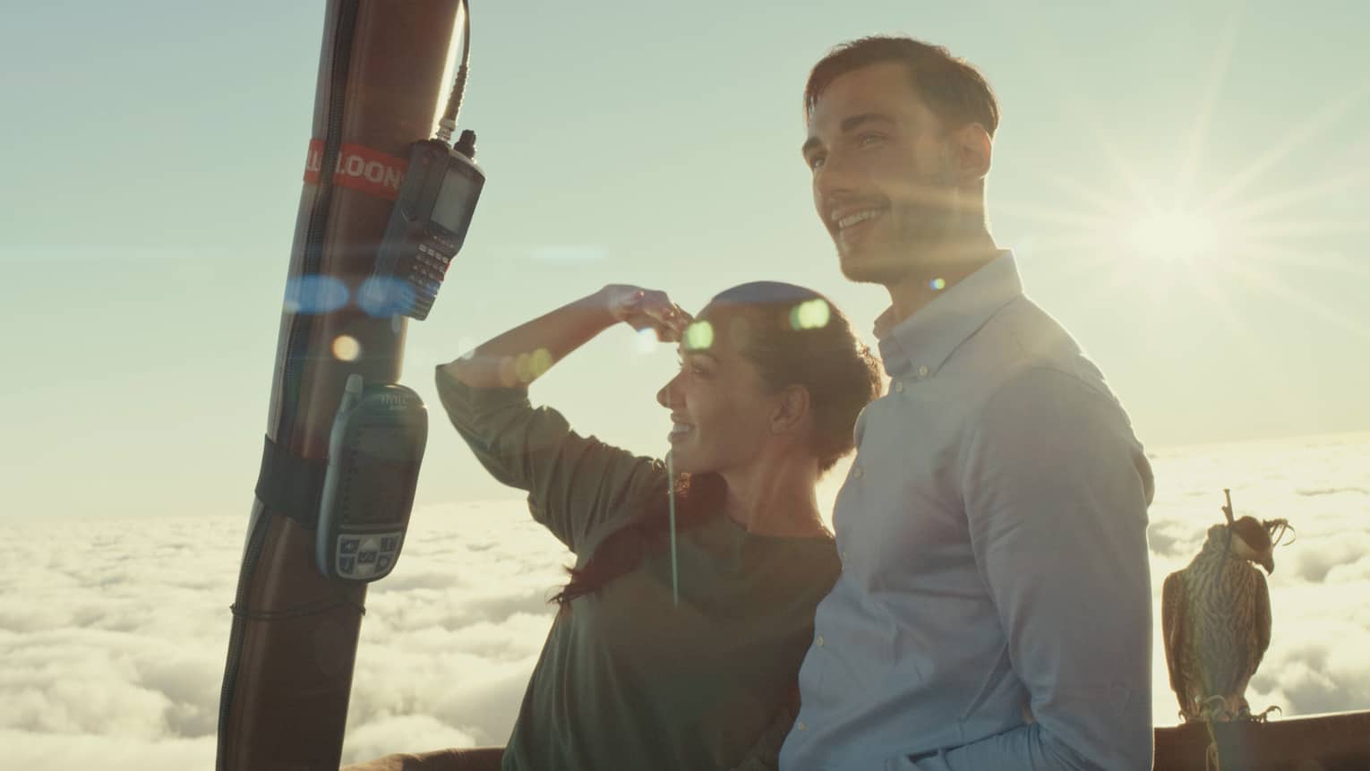 Two guests high above the clouds in a hot air balloon, gazing ahead; a hooded falcon perched on the railing behind them.