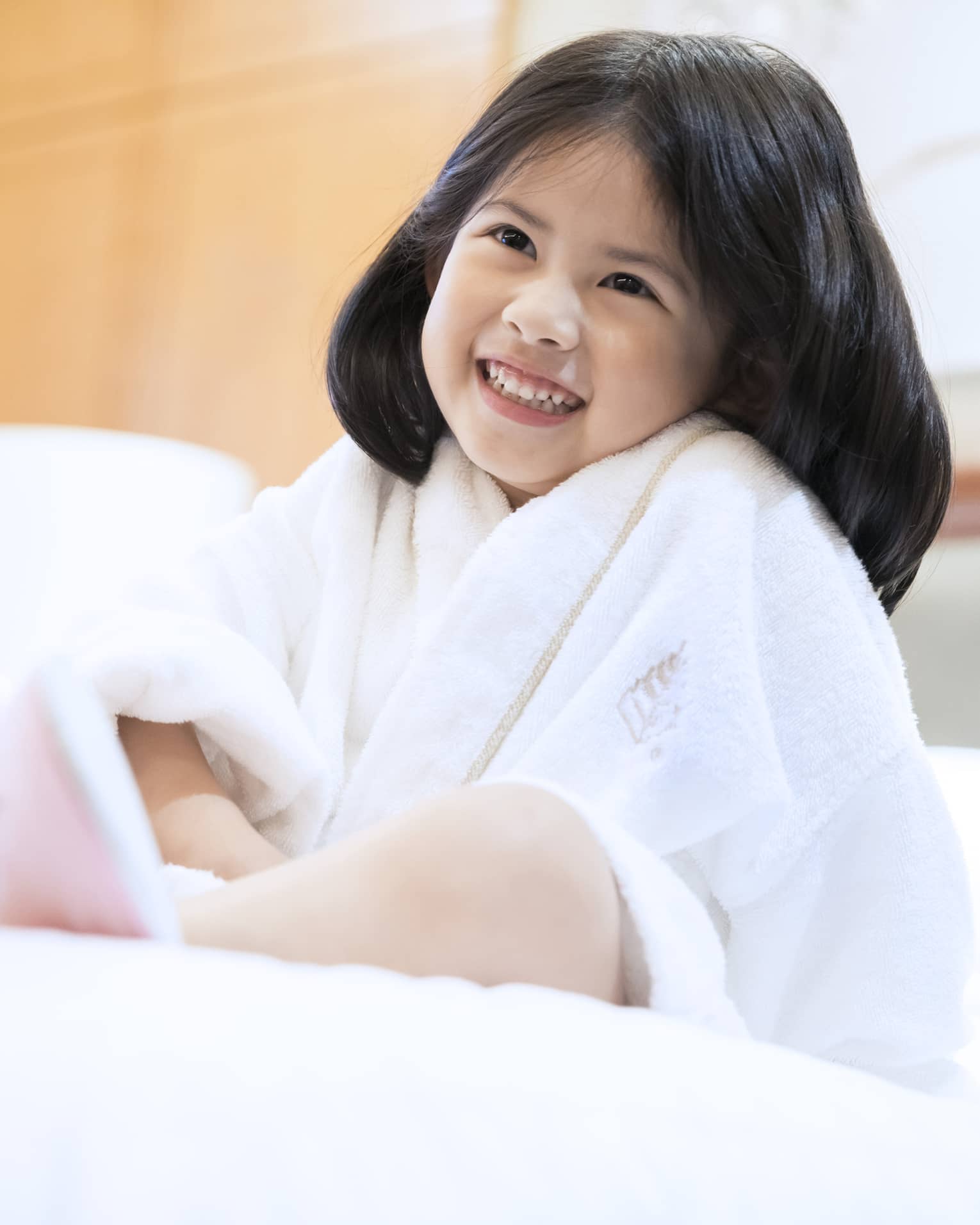 Smiling little girl wearing white bath robe and pink slippers sits on bed