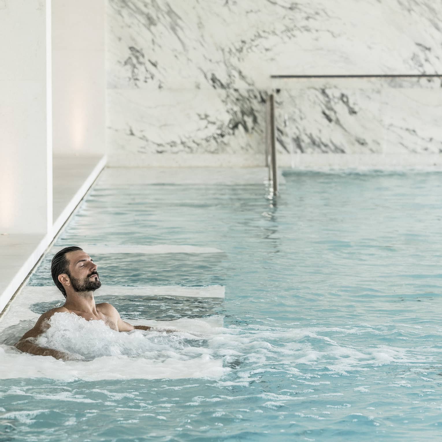 Man with beard relaxes in spa waters, surrounded by white marble