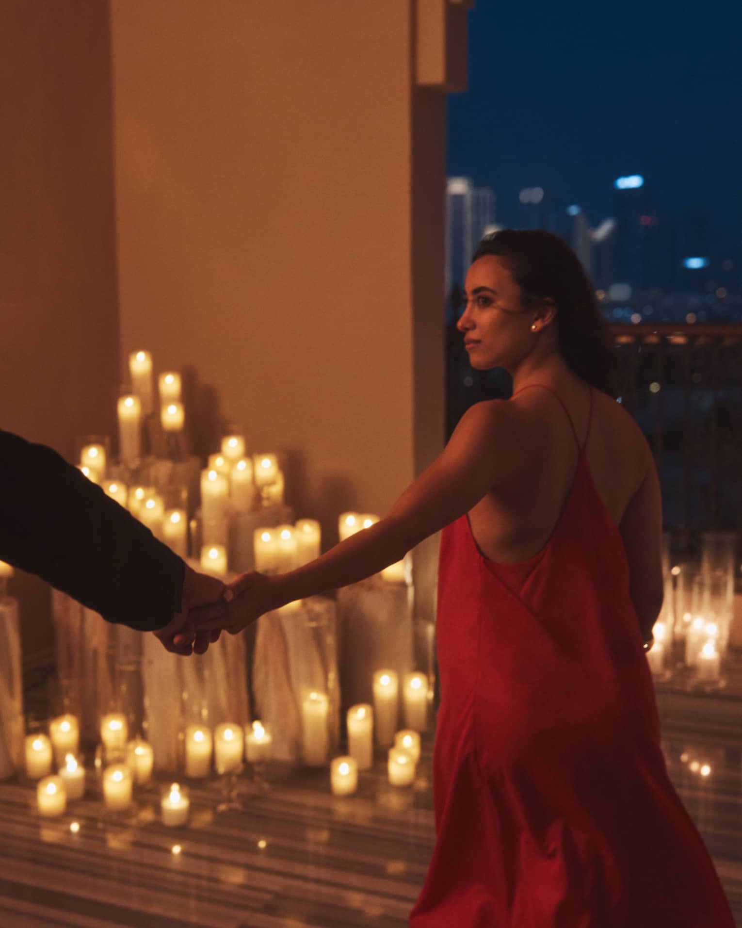 Person wearing a red dress leads another person who is silhouetted in the shadow by the hand toward a terrace filled with lit candles at night