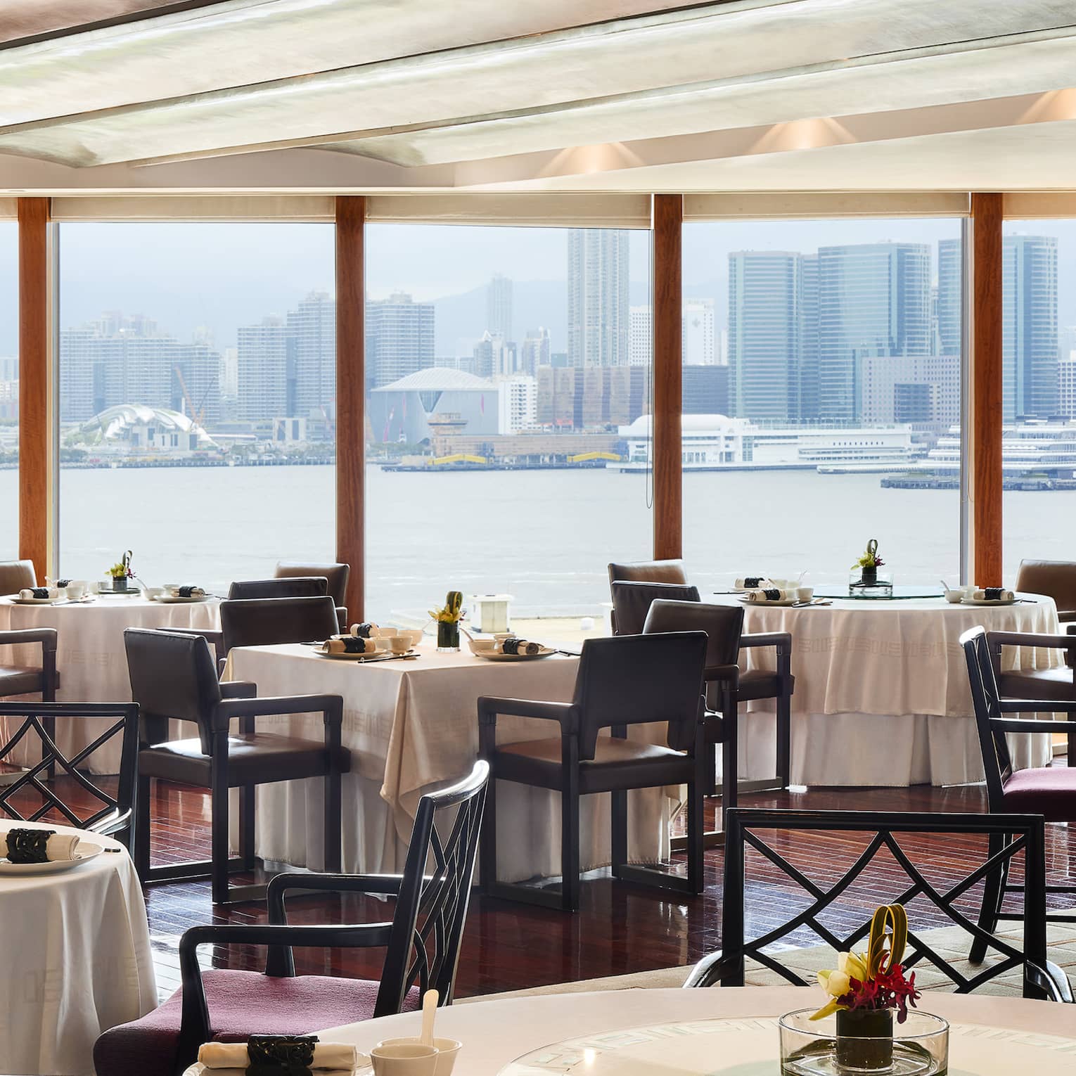 An elegant dining room with tables set with flowers and place settings, overlooking the city from across a body of water.