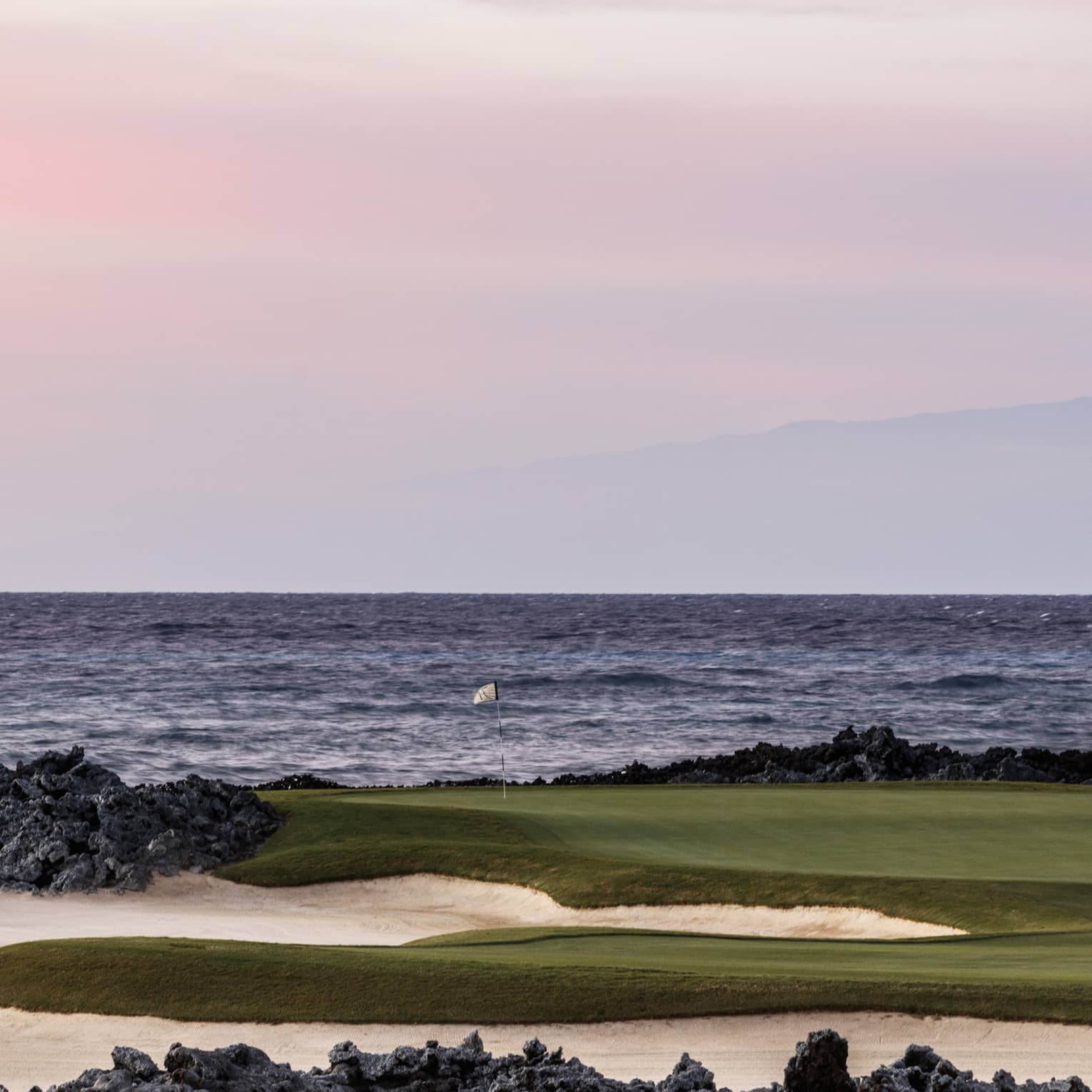 On an oceanside golf course beneath a rose-coloured sky, a single flagstick stands amid black lava rocks and white sand. 