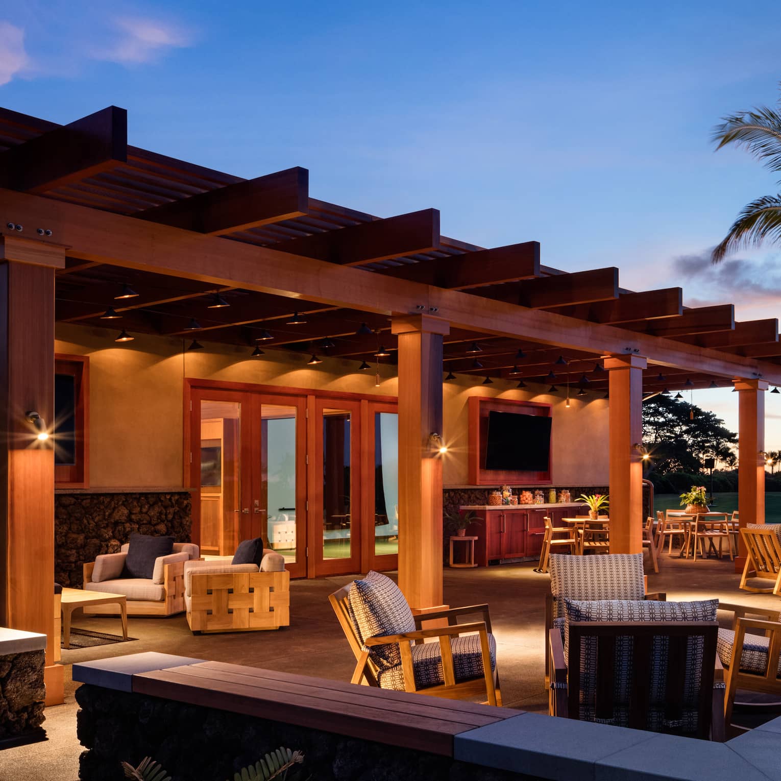 A warmly lit patio furnished with tables and cushioned armchairs under a clear, royal-blue night sky, sunset in the distance.