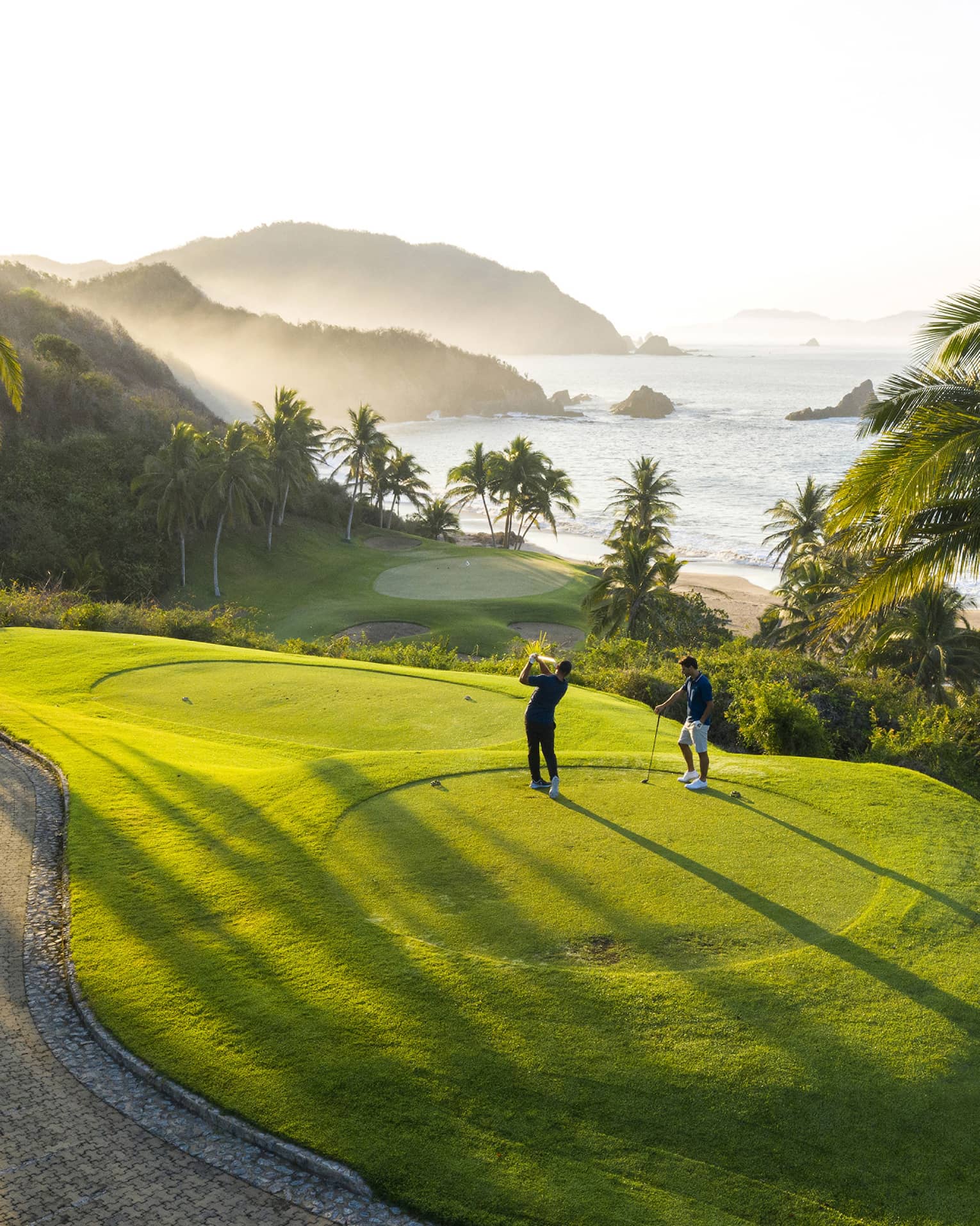 Rear view of two golfers on an elevated tee, one swinging toward the green down by the beach, misty hills and ocean beyond.