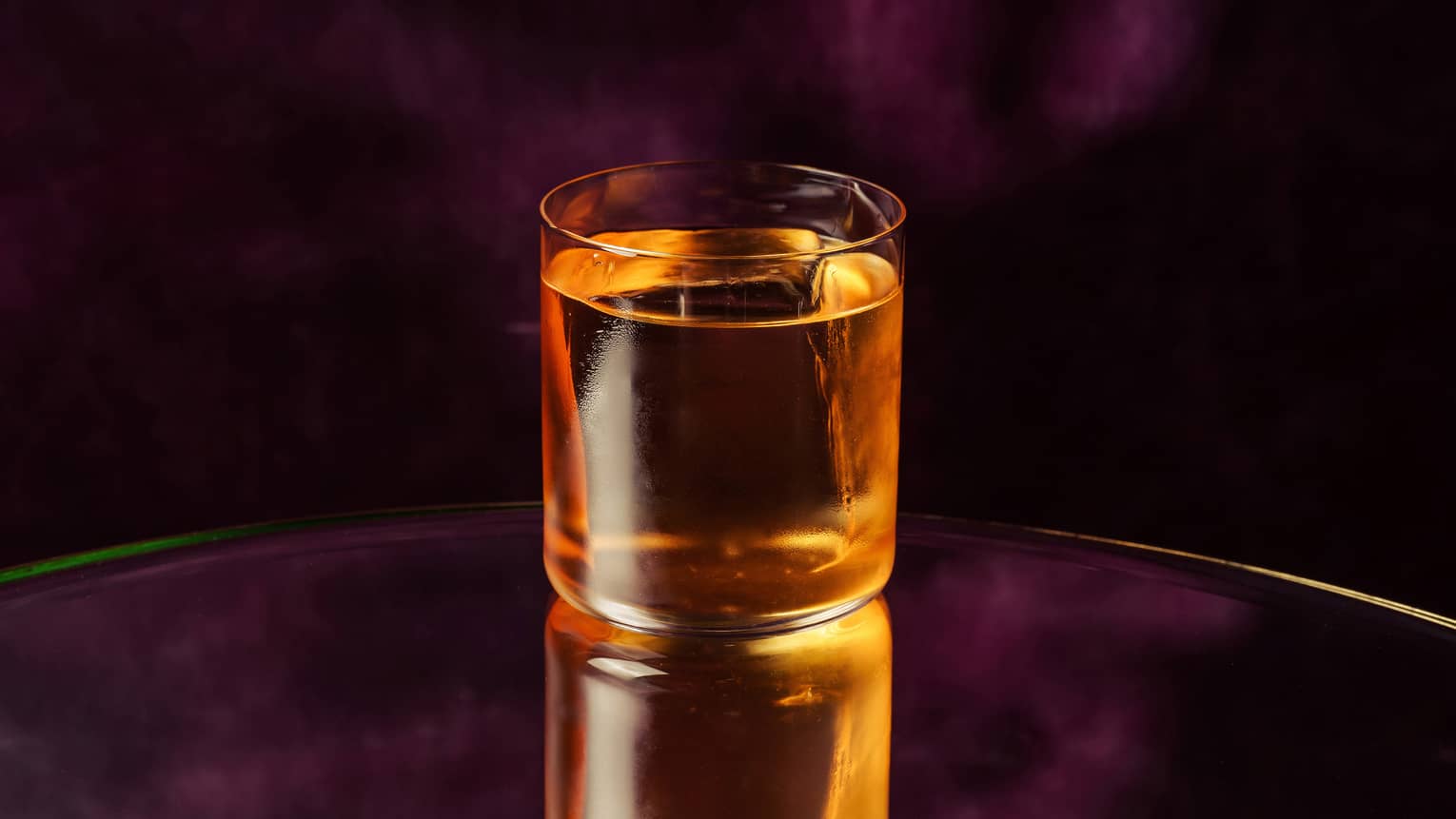 A golden-colored beverage in a rocks glass shown on a dark violet backdrop
