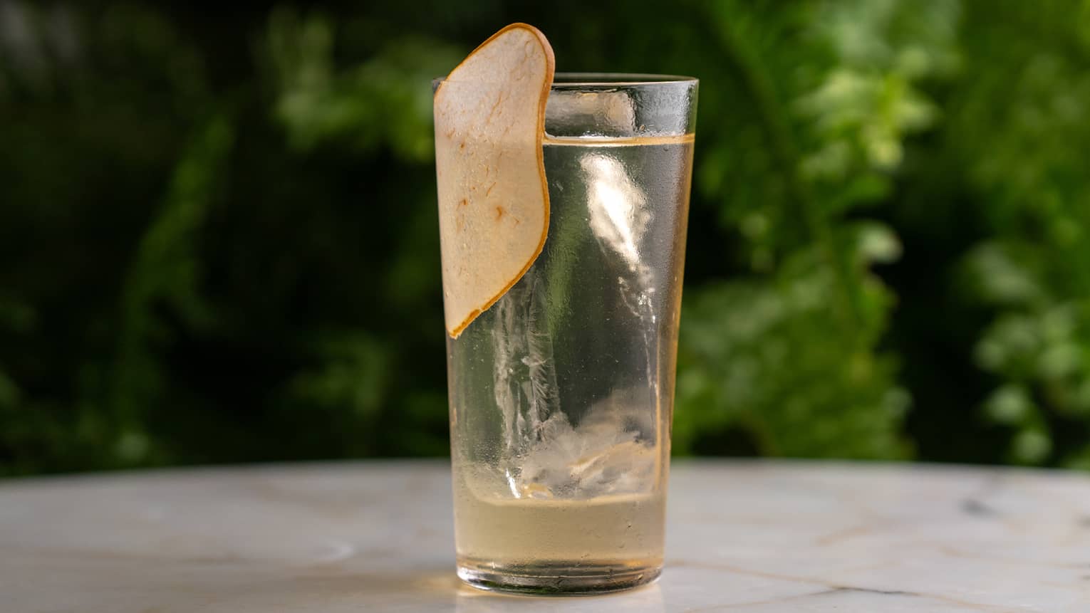 A tall tumbler holds a transparent light peach-colored cocktail with garnish.