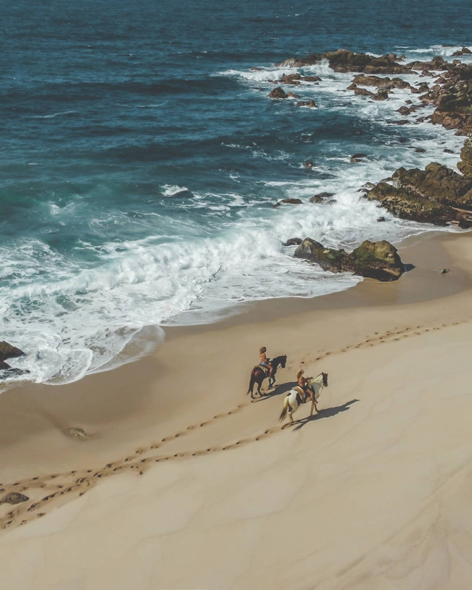 Two people riding horses on the beach next to the ocean.