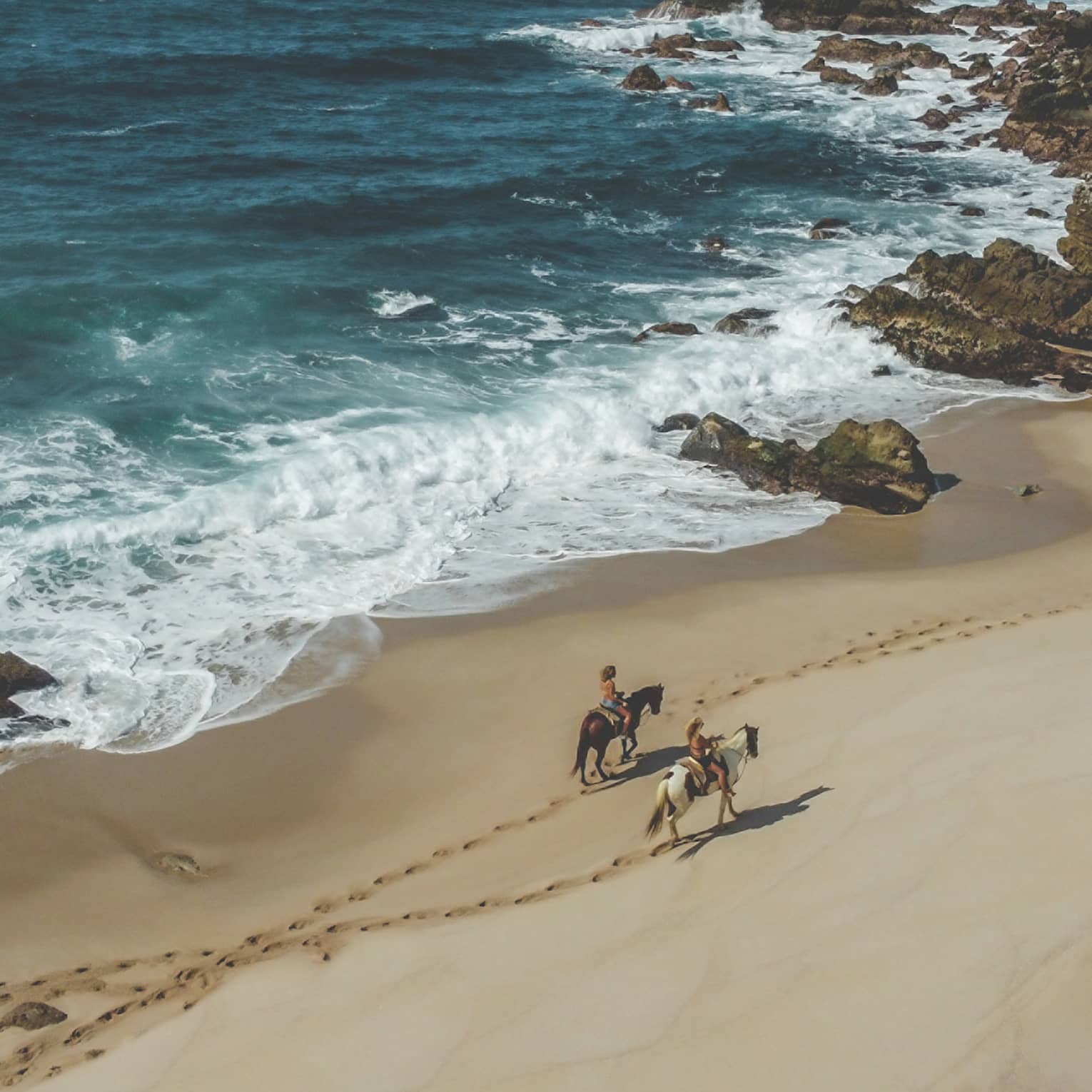 Two people riding horses on the beach next to the ocean.