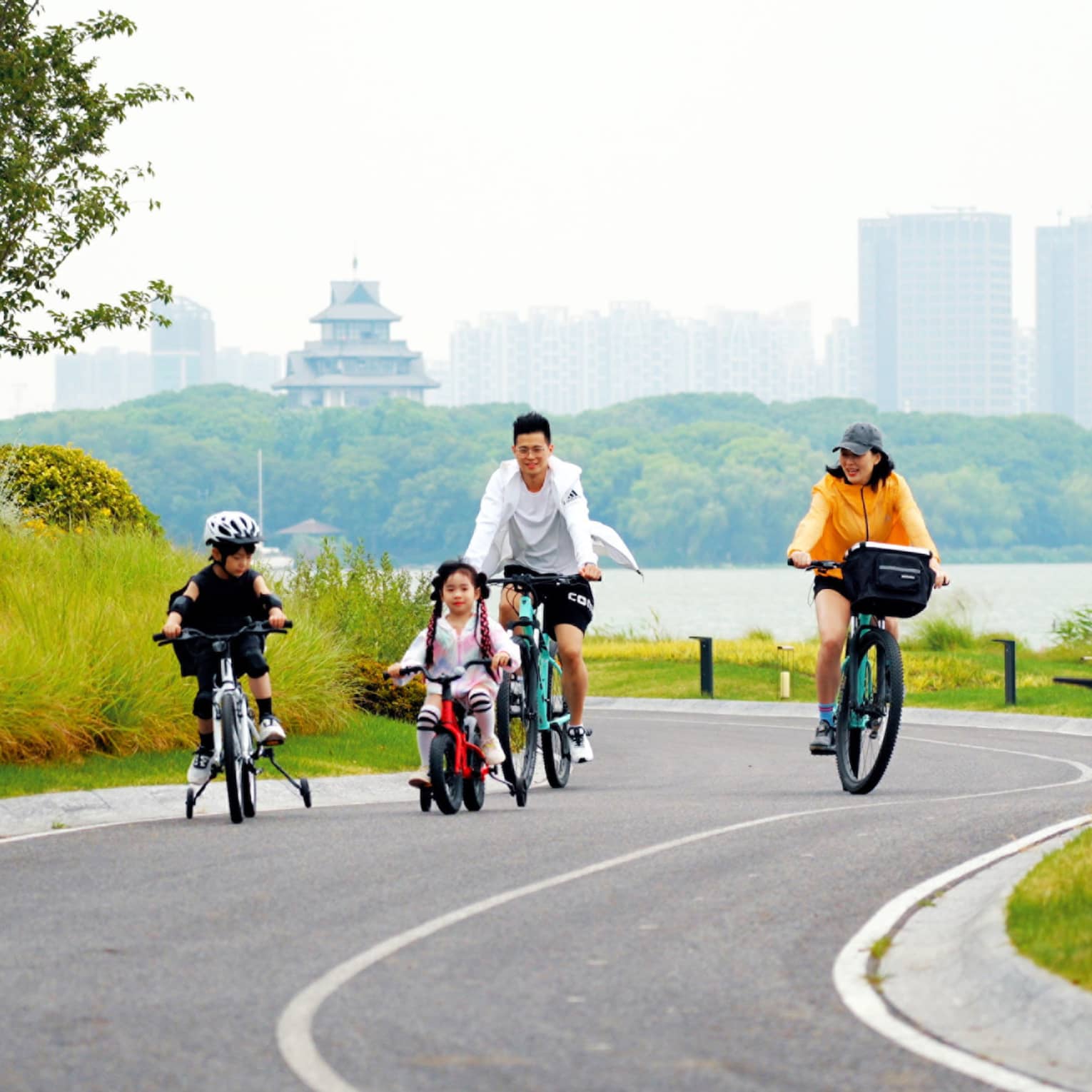 A family cycles uphill on a winding road bordered by bright grass and foliage, a hazy lake, forest and cityscape behind them.