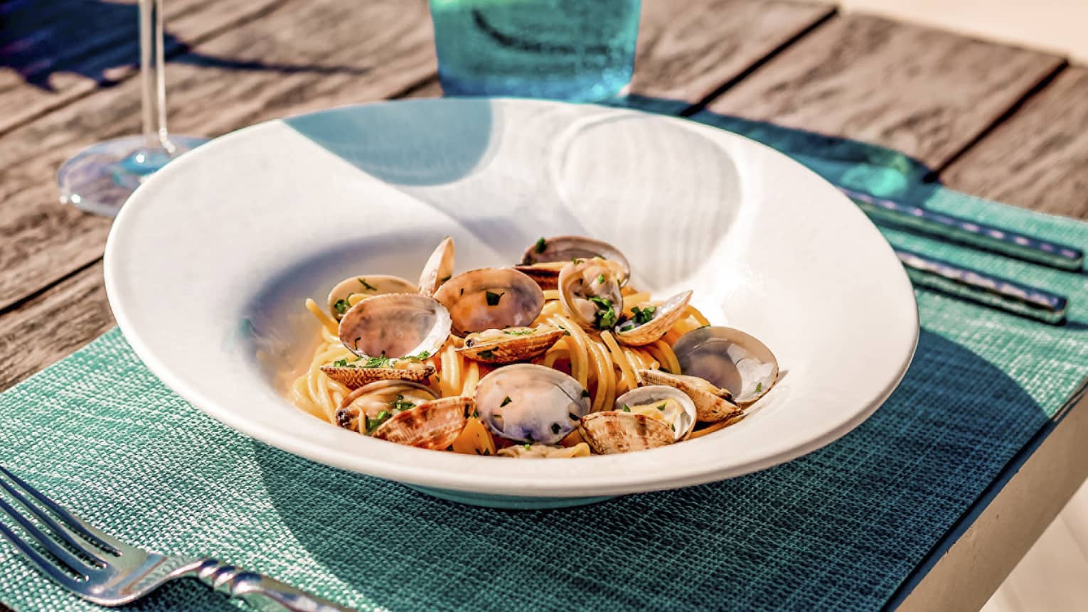 Spaghetti alle vongole on wooden picnic table with turquoise placemat and water glass, clear wine glass, conch shell