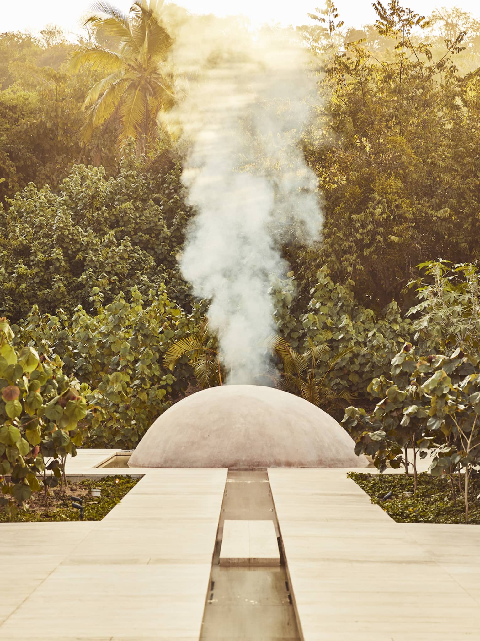 White steam rises from a small cement dome nestled between two large stone walls and surrounded by dense dark-green foliage.