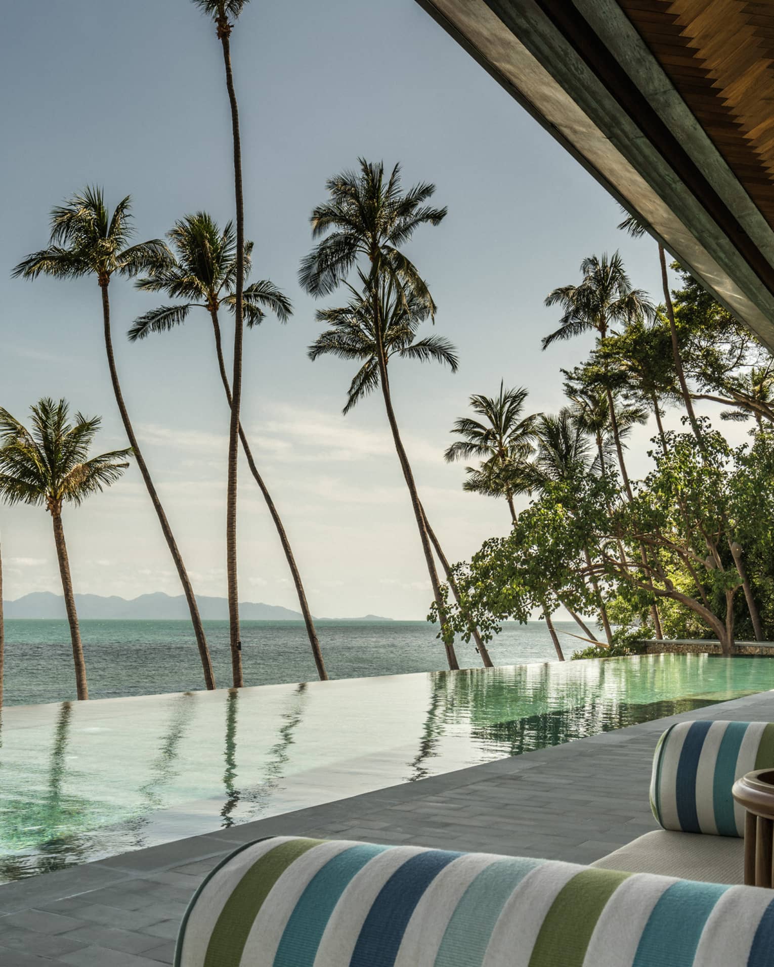 Green-and-blue striped banquettes on patio by infinity swimming pool, ocean