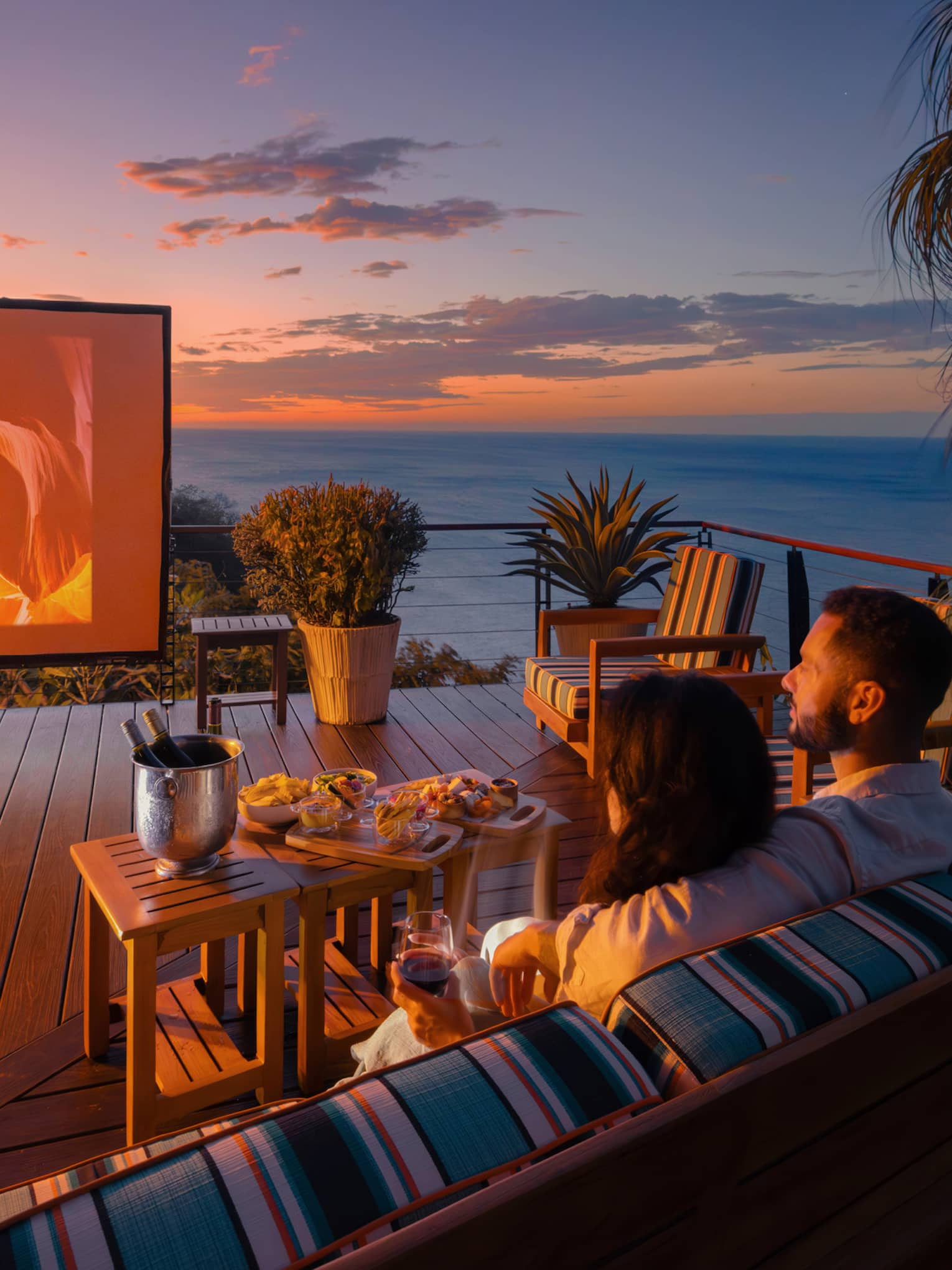 Two people sit on an outdoor sofa with a small table of food infront of them as they watch a movie on a large outdoor screen set up on a private terrace