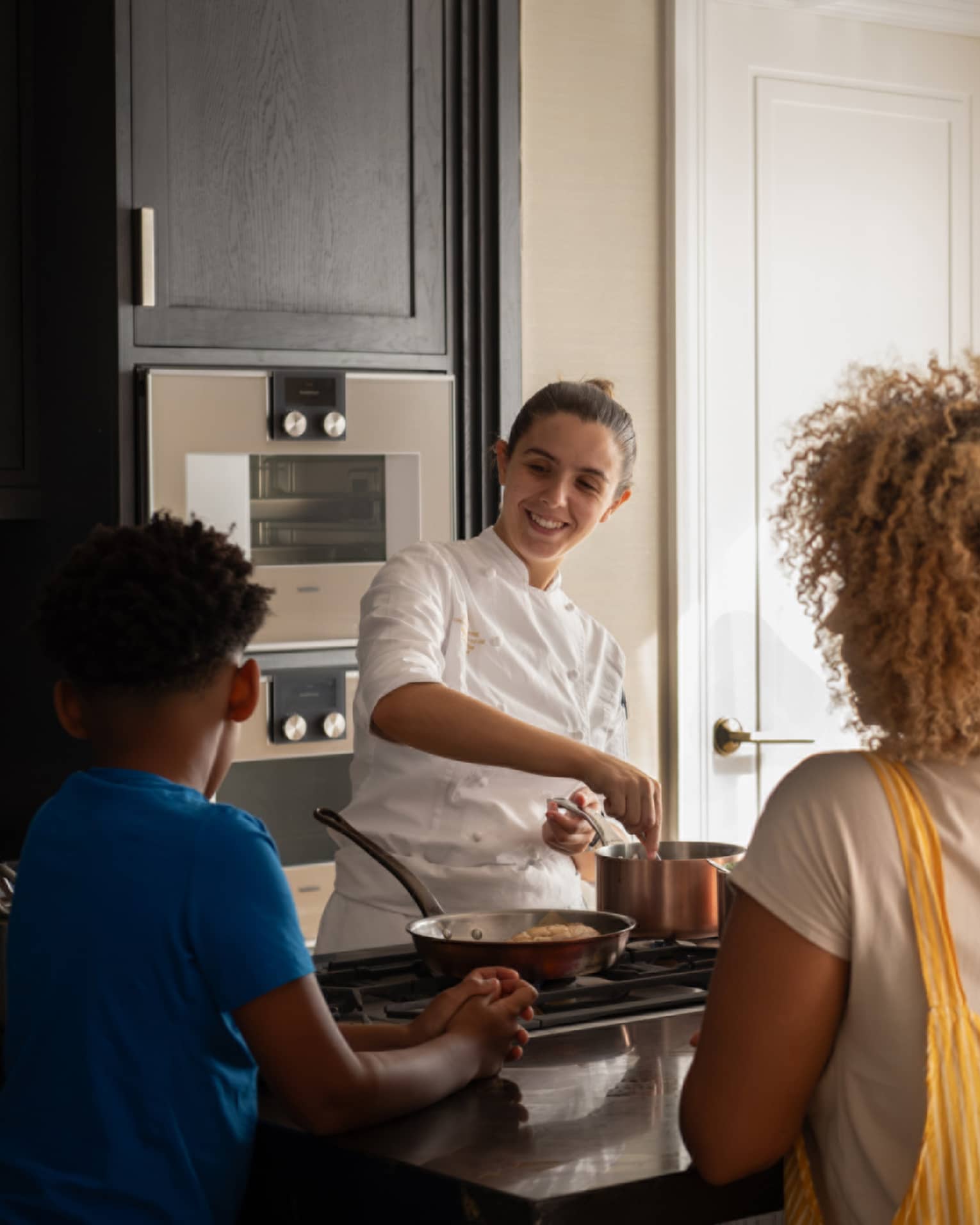 A family of four gathers around a kitchen-island cooktop watching a smiling chef prepare food in copper cookware. 