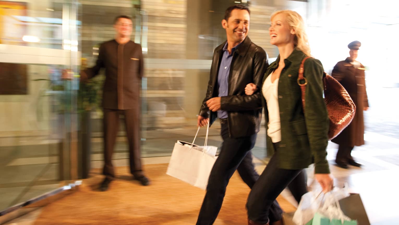 Man and woman wearing black coats and carrying shopping bags walk arm-in-arm into hotel lobby