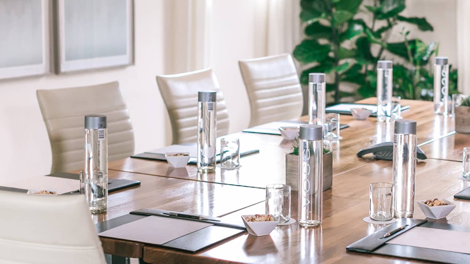 A rectangular boardroom table set with clear glass water bottles and black folders in a naturally-lit room
