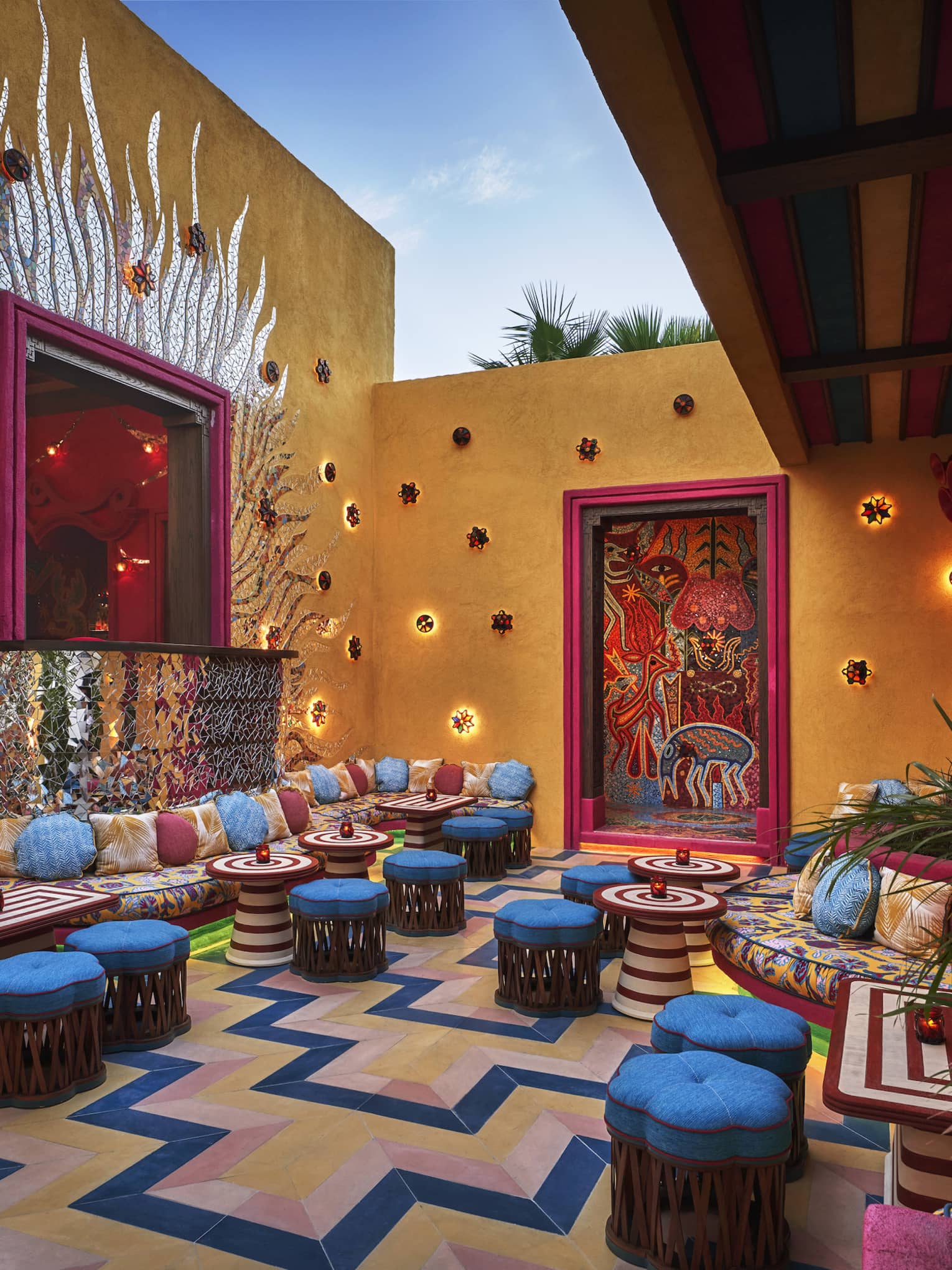 A colourful outdoor seating area with bright blue cushioned stools, colourful lights and plants.